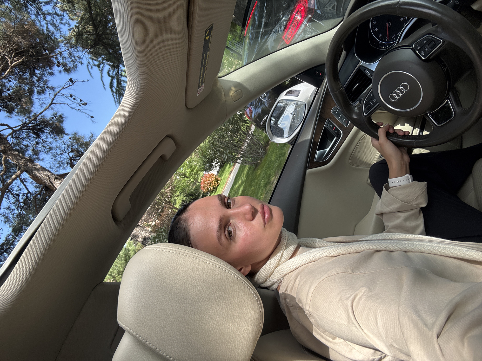 A woman reclines in the driver's seat of an Audi, head resting on the headrest, hand on the steering wheel. She wears a beige jacket and a white scarf. The car is parked, and trees and a building can be seen through the windows. The location is likely Tbilisi, Georgia, based on the provided instructions.
