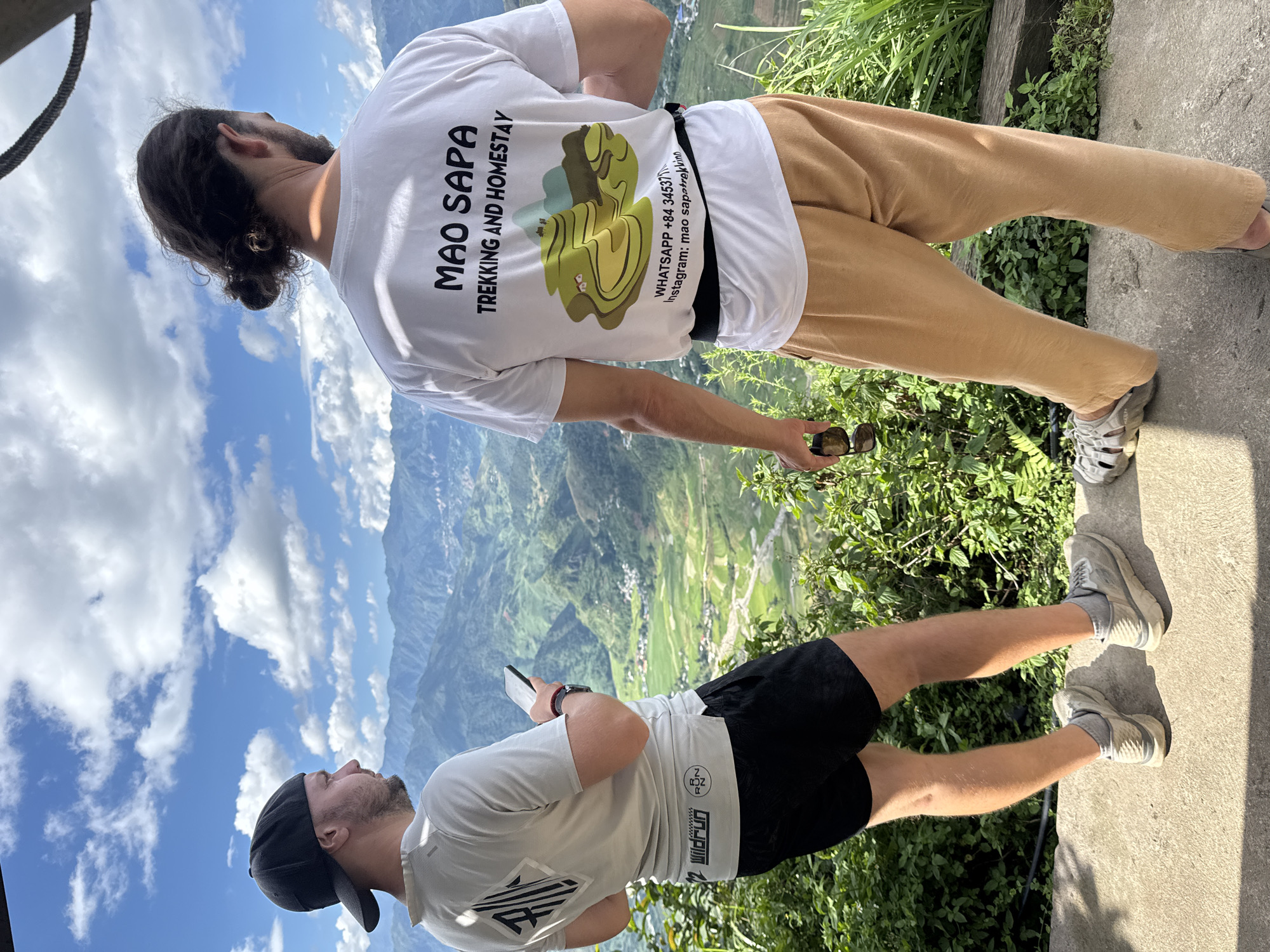 Two men stand on a concrete overlook, gazing at a mountainous valley in Tu Le, Vietnam, under a blue sky with scattered white clouds.
The man on the left wears a black baseball cap backward, a light grey t-shirt with a dark geometric design ("RZN") and "Wildrun" text, black shorts, and light grey athletic shoes. He holds a black smartphone in his right hand and looks towards the left side of the frame.
The man on the right, viewed from behind, has dark hair tied in a bun and a short beard. He wears a white t-shirt featuring "MAO SAPA TREKKING AND HOMESTAY" in black text, an illustration of green terraced mountains with small houses, and contact information including "WHATSAPP: +84 34537..." and "Instagram: mao_sapatrekking". He also wears light brown, loose-fitting pants, grey open-toed sandals, and a black waist belt. He holds dark sunglasses in his right hand and looks out over the valley.
The background reveals a vast, lush green valley characterized by extensive terraced fields, indicative of rice paddies, and small settlements nestled among numerous layered mountains. Green foliage grows along the edge of the concrete platform in the immediate foreground. A dark rope or cable is visible in the upper right corner.