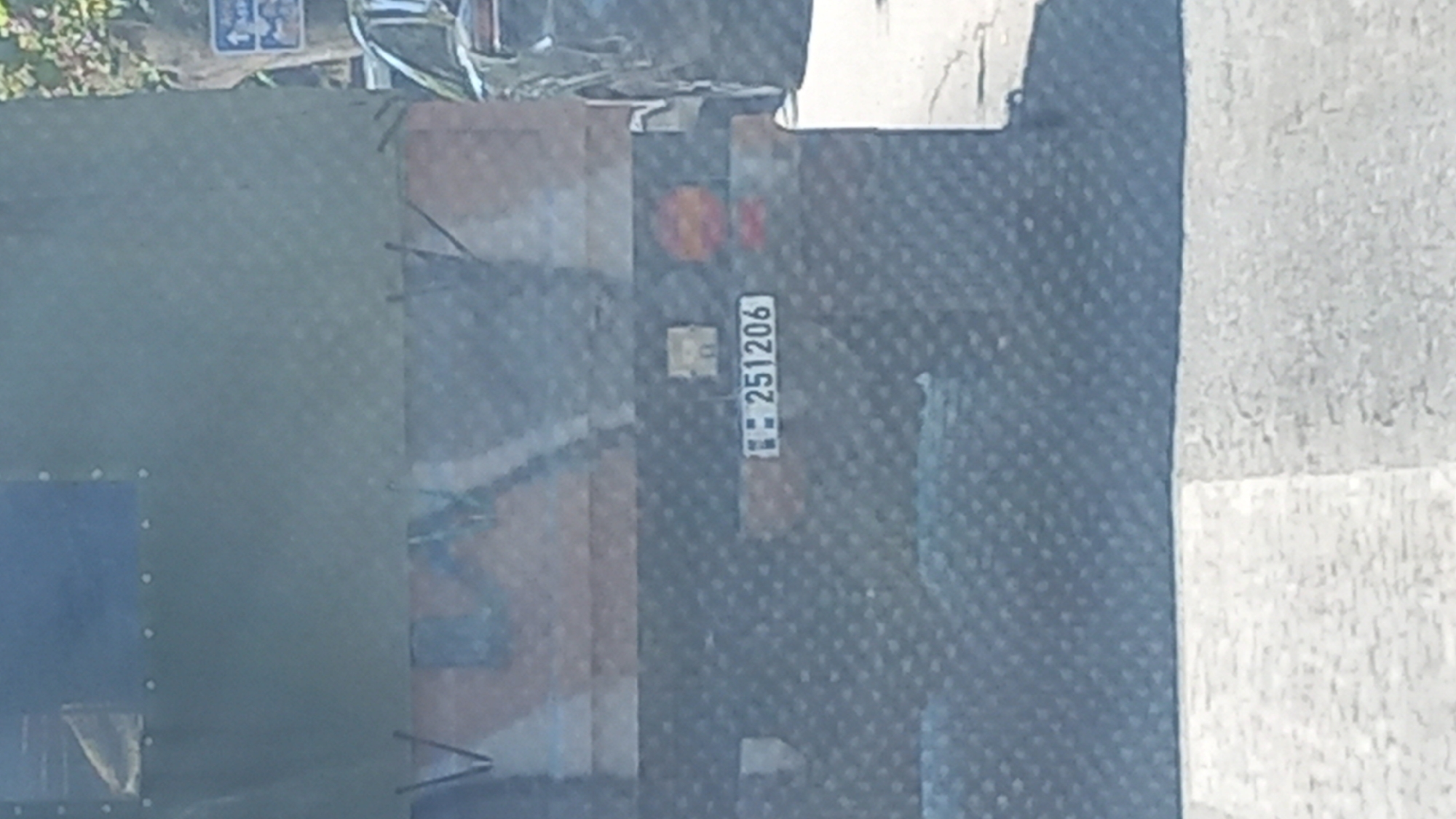 The image is a close-up, slightly blurry, outdoor photograph of the rear of a military-style truck. The truck has a dark, textured rear panel and a tarpaulin covering the top, which is olive green. A license plate with the number "251206" is visible, along with what appear to be taillights. To the right of the truck, the rear of a black SUV is partially visible. The scene appears to be on a paved road, and in the background, trees and a blue directional sign can be seen, suggesting an urban or suburban environment. The lighting indicates it is daytime. The location is identified as Kos, Greece.