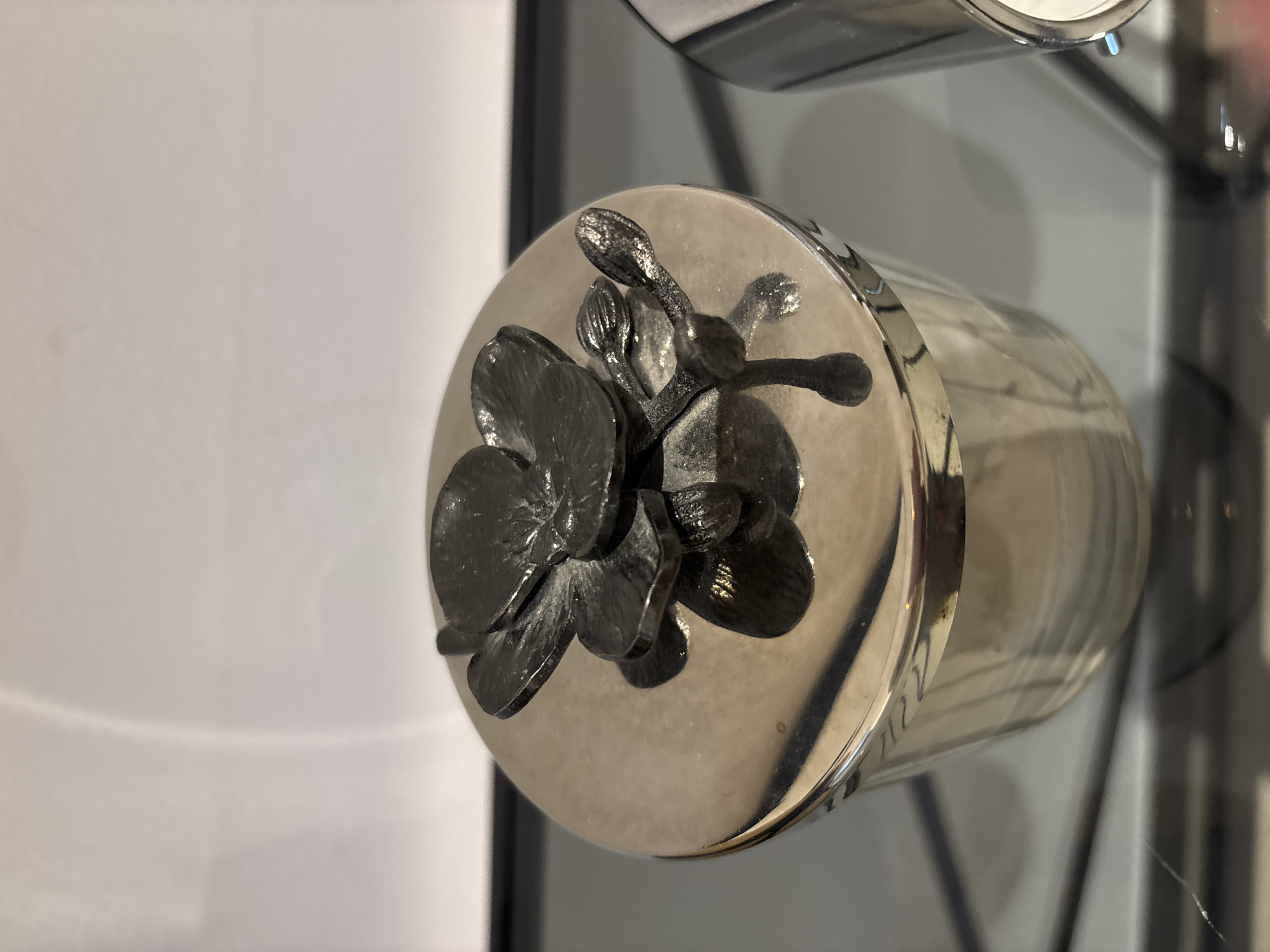 A close-up, high-angle shot shows the lid of a shiny silver jar, adorned with a dark, metallic orchid decoration. The jar itself appears to be made of clear glass, with the contents inside not visible. The lid is circular and has a reflective surface. The orchid is intricately detailed, with multiple petals and smaller bud-like elements. The background is a soft, out-of-focus white surface, suggesting an indoor setting. The scene is clean and well-lit, with a slight shadow cast by the orchid onto the lid. The image is sharp and focused on the jar and its embellishment. The object is likely in a retail display or home decor setting within Dubai, United Arab Emirates, given the context of the request.