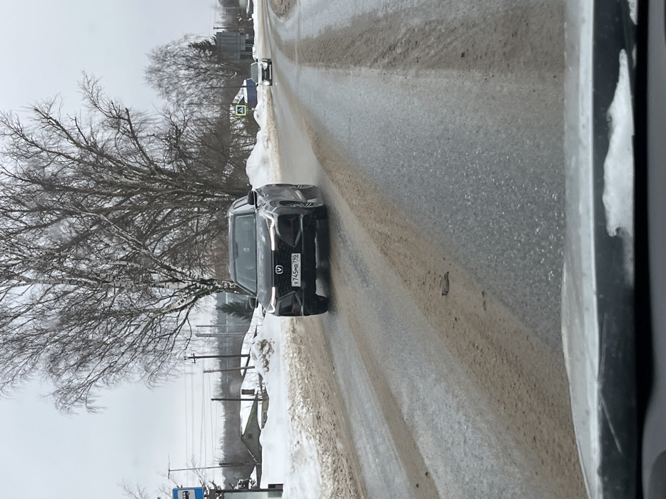 The scene depicts a grey winter day on a two-lane paved road in Tatishchevo, Russia. A dark grey or black Changan SUV, bearing Russian license plate X745MB 799, drives towards the viewer in the left lane. Its daytime running lights are illuminated. The road surface is wet, showing streaks of melted snow, mud, and slush, with compacted snow and dirt visible on the shoulders. Bare deciduous trees line both sides of the road, and in the background to the left, several single-story buildings are visible amidst snow-covered ground. A blue bus stop sign is present on the left side of the road. Further down the road on the right, a white or silver sedan is visible driving away from the viewer, near a pedestrian crossing sign. The image appears to be taken from inside another vehicle, with the top of its dashboard visible at the bottom of the frame. No individuals are explicitly visible.