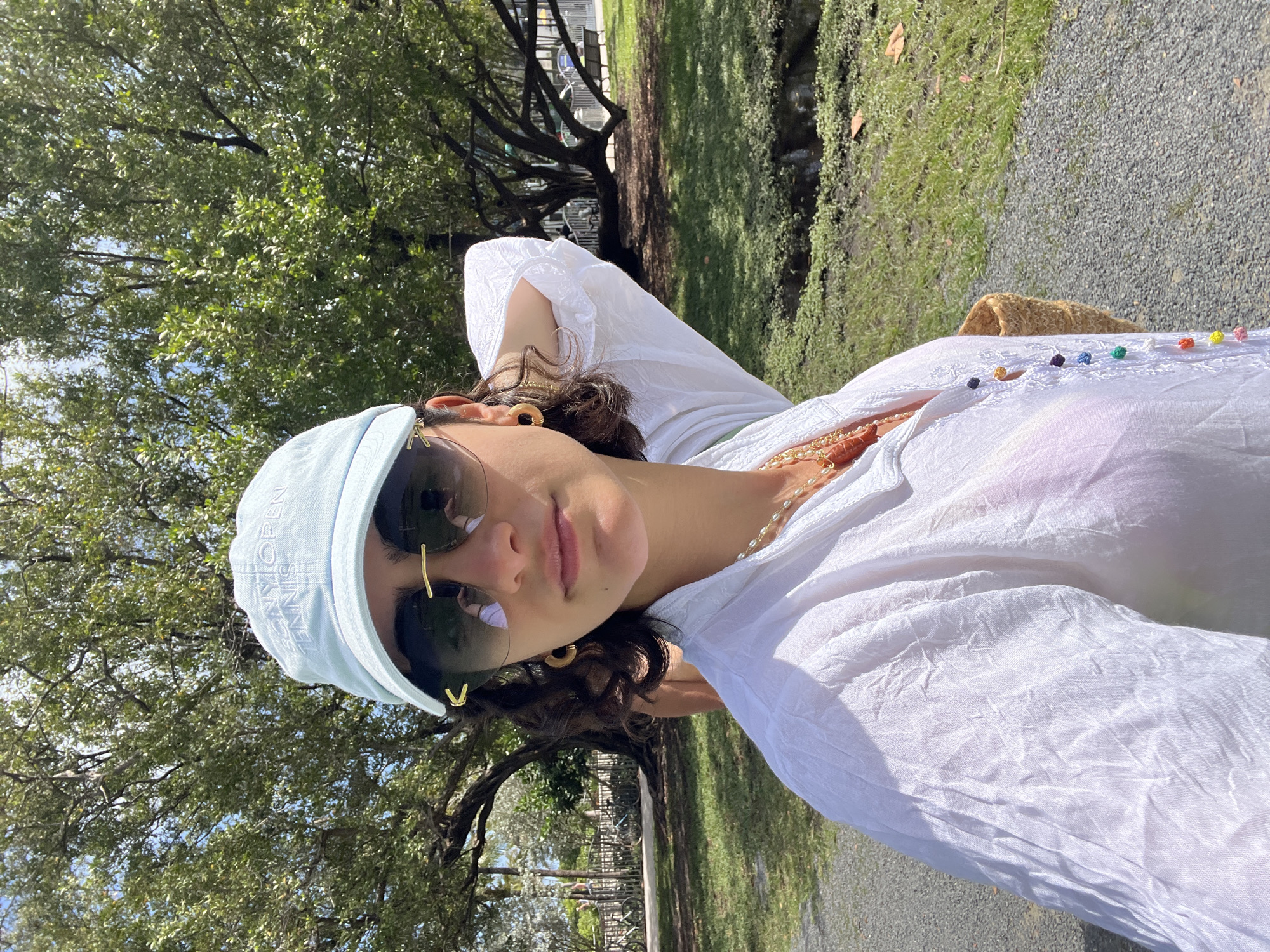 A young adult female in a white shirt, light blue cap and sunglasses is captured in an outdoor shot, likely taken in Miami Beach, United States of America. The background features foliage, likely a park setting with trees and greenery. Some park fixtures are visible behind the subject. The woman is positioned in the foreground, angled towards the camera.