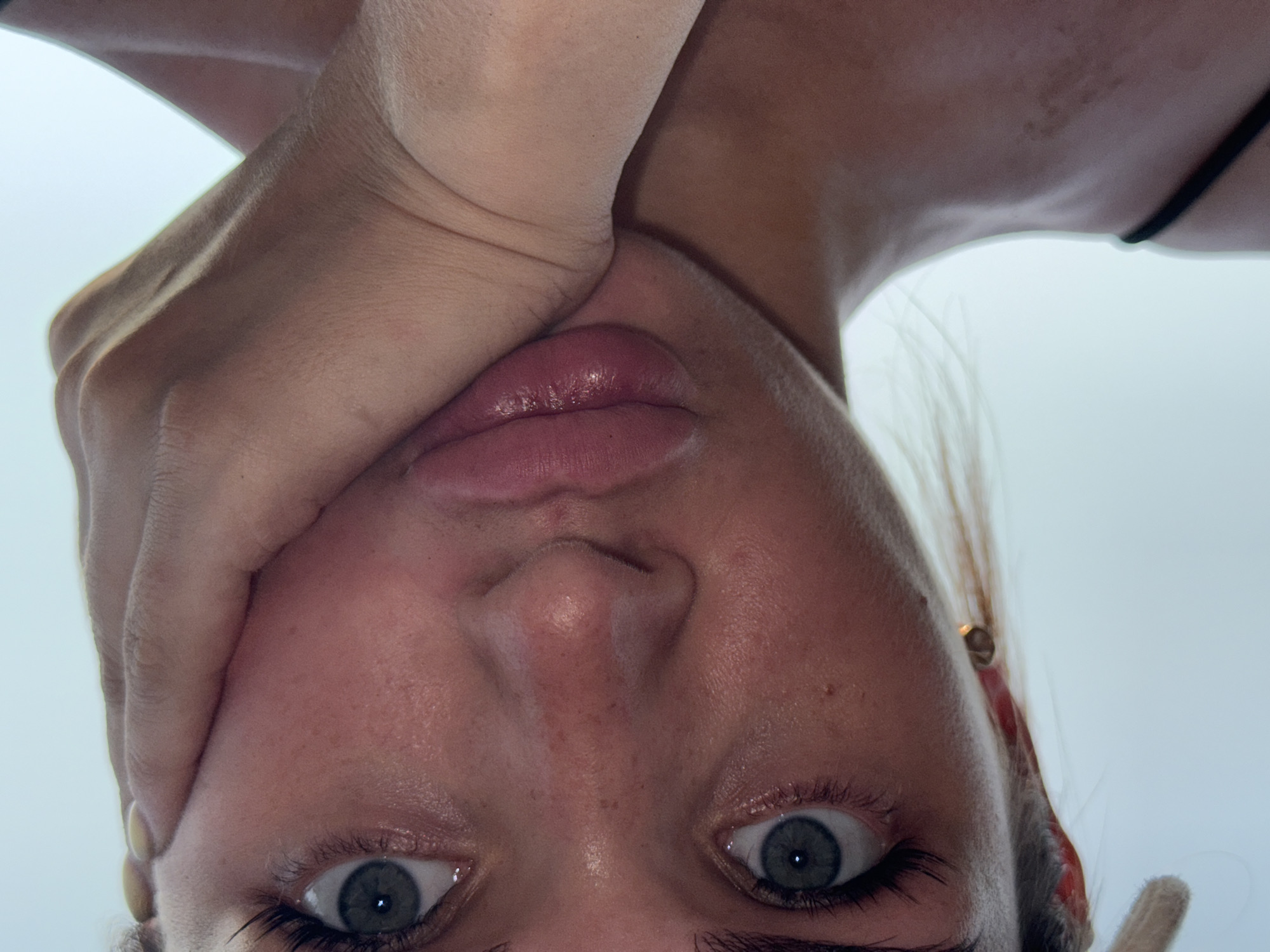The image is a close-up, low-angle shot of a woman's face and upper torso, set against a bright, hazy sky. She has light skin with visible freckles and a slightly flushed complexion. Her eyes are open, with striking blue irises and dark eyelashes. Her lips are full and appear to be slightly pursed, with a sheen of gloss. One hand is resting against her cheek, with her fingers extended upwards towards her eye. She is wearing a black strap, likely from a tank top or dress, visible on her left shoulder. The background is a uniform, pale blue, suggesting an outdoor setting, possibly during daylight. There are no discernible features that place this image in the city of Лобаново, Russia; the environment appears generic and devoid of specific urban indicators.