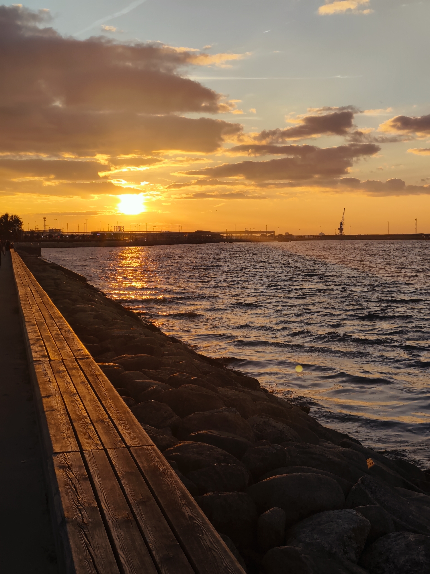 The image depicts a scenic sunset over the Baltic Sea at the Tallinn Seaplane Harbour. In the foreground, a weathered wooden walkway runs along the left side, bathed in the warm, golden light of the setting sun. To the right of the walkway, a rocky embankment slopes down to the water. The sea is choppy, with ripples reflecting the vibrant orange and yellow hues of the sky. In the mid-ground, a pier extends into the water, lined with lamp posts. Further in the distance, industrial cranes and structures of the port are silhouetted against the dramatic sunset. The sky is filled with a mix of wispy clouds tinged with orange and pink, and areas of clear blue. A thin contrail is visible in the upper part of the sky. The overall scene is one of natural beauty enhanced by the industrial landscape of the harbor.