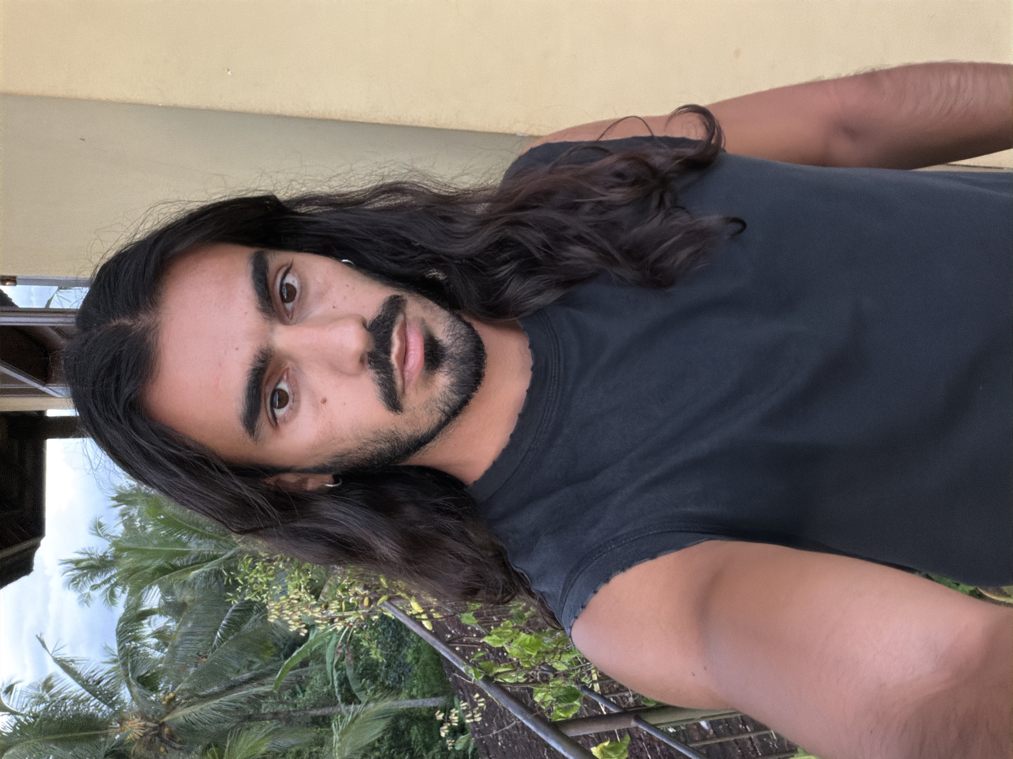 A close-up selfie of a man with long, dark, wavy hair and a beard, wearing a black sleeveless shirt. He has a light complexion and dark eyes. Behind him, lush green palm trees and foliage are visible, suggesting a tropical outdoor setting. A yellow wall and a railing are also present in the background, consistent with a balcony or terrace in Tegallalang, Indonesia, a location known for its rice terraces and natural beauty. The lighting indicates it is daytime, possibly late afternoon or early evening.
