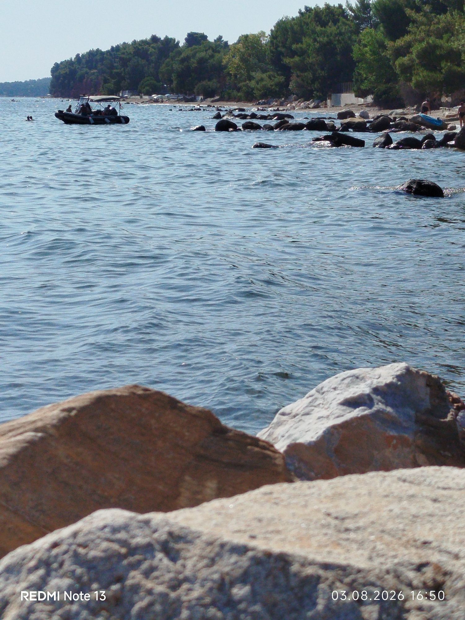 A wide shot captures the shoreline of Sithonia Municipal Unit, Greece, under a clear sky. The foreground is dominated by large, textured rocks, one reddish-brown and the other a lighter gray. Beyond these rocks, the calm, blue sea stretches towards a tree-lined coast. A dark inflatable boat with a canopy carrying several people navigates the water in the mid-ground, with two individuals visible swimming further to the left. The coastline is dotted with more rocks and a few scattered beach umbrellas. The image is dated "03.08.2026 16:50" and branded "REDMI Note 13".