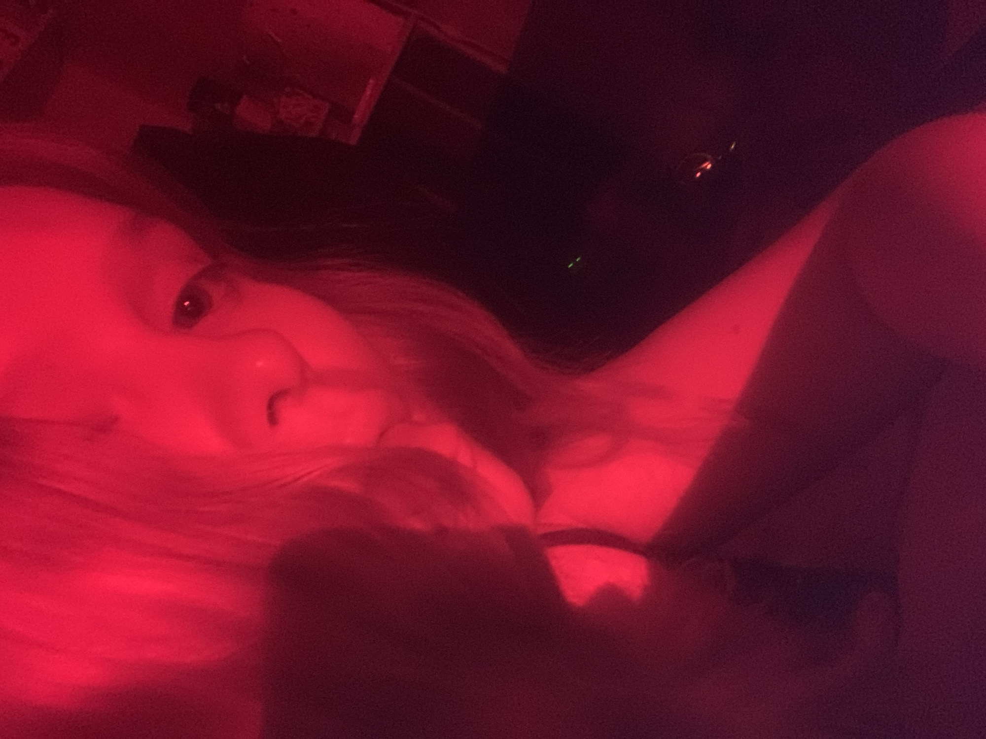 The image captures a person, likely female, in a close-up shot dominated by intense red lighting. Her head and upper torso are visible, with her face angled towards the viewer. Her left eye, appearing dark, looks directly at the camera, while her nose and mouth area are partially obscured, possibly by her hand or arm. Long hair, saturated in red light, frames her face and extends around her.
The subject appears to be reclining or lying down in an intimate, low-light indoor setting, possibly a bedroom. A dark strap, likely from clothing, is visible on her right shoulder/chest area. The background is largely indistinct and dark, with a few small, glowing points of light – some green and others vaguely red/orange – suggesting the presence of electronic devices or ambient indicators. The strong red illumination creates a dramatic, monochromatic mood, making it difficult to discern natural colors of objects or clothing. No visible text is present in the image.