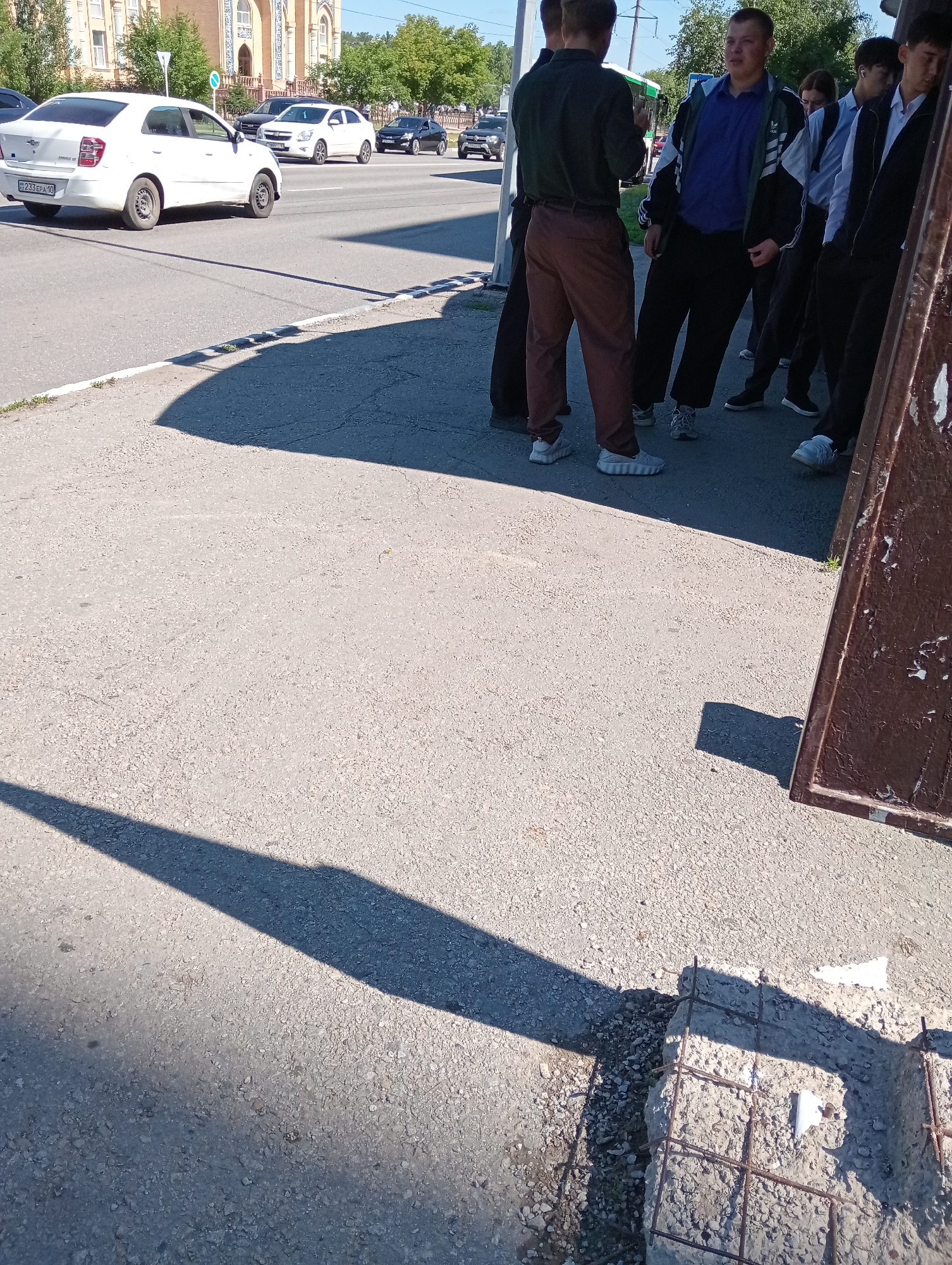 The image shows a group of young men standing on a sidewalk next to a street in an urban environment. They appear to be in casual attire, with some wearing jackets and pants, and others in shirts and pants. The men are standing or leaning against a brown structure, possibly a fence or a large signpost, and their shadows are cast on the pavement. In the background, there are cars on the street, including a white sedan and a black sedan. Buildings and trees are also visible in the distance. The ground is paved with asphalt, and there are some patches of grass and debris along the edge of the sidewalk. The lighting suggests it is daytime, with the sun casting strong shadows.