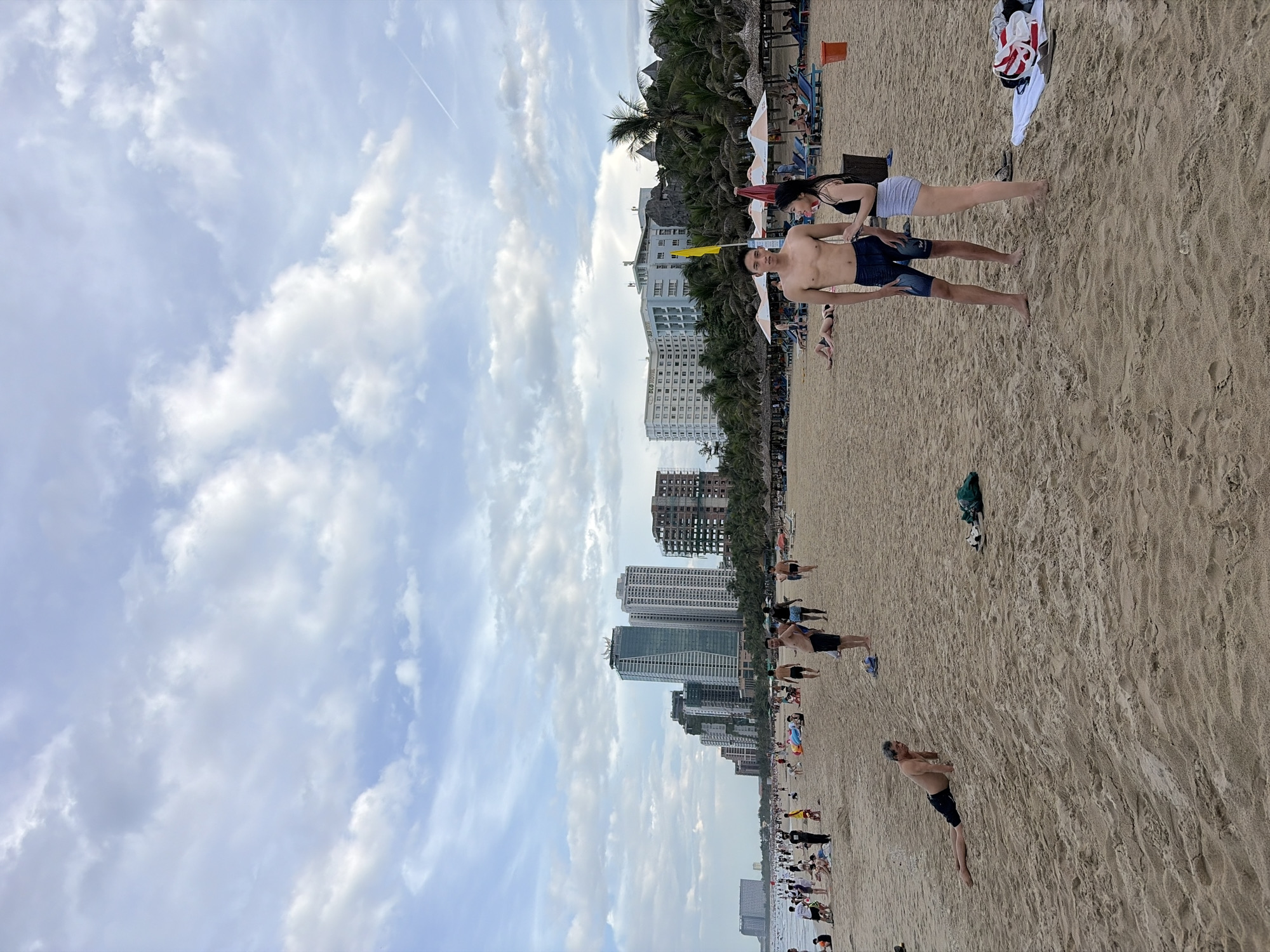 This image captures a crowded beach scene in Đà Nẵng, Vietnam. In the foreground, two young adults, a shirtless man in denim shorts and a woman in a black tank top and grey shorts, are standing on the sand. To the left, an older man is doing push-ups on the beach. Scattered across the sand are beachgoers, some sitting, some walking, and some sunbathing. In the background, a row of tall buildings, including hotels and apartments, lines the coastline, with palm trees interspersed among them. The sky is partly cloudy with a light blue hue. Beach umbrellas and lounge chairs are visible further down the beach, indicating a popular tourist area.