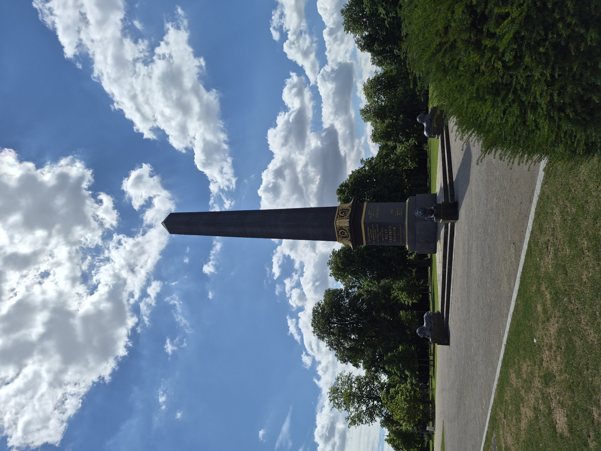 This image, taken in Brunswick, Germany, showcases a monument outdoors on a sunny day with scattered clouds. The monument is a tall, dark obelisk with an ornate, golden base featuring inscriptions. The text on the base is partially visible, including phrases like "DES VATERLANDES" (Of the Fatherland) and names such as "WELFE FRIEDRICH WILHELM".
Flanking the monument are dark, stylized lion sculptures. The monument and sculptures are set on a paved pathway, with neatly manicured green lawns on either side. In the background, a line of lush green trees creates a natural backdrop. The sky is a vibrant blue, punctuated by fluffy white clouds, suggesting pleasant weather, likely daytime. The overall scene conveys a sense of solemnity and historical significance, befitting a monument.