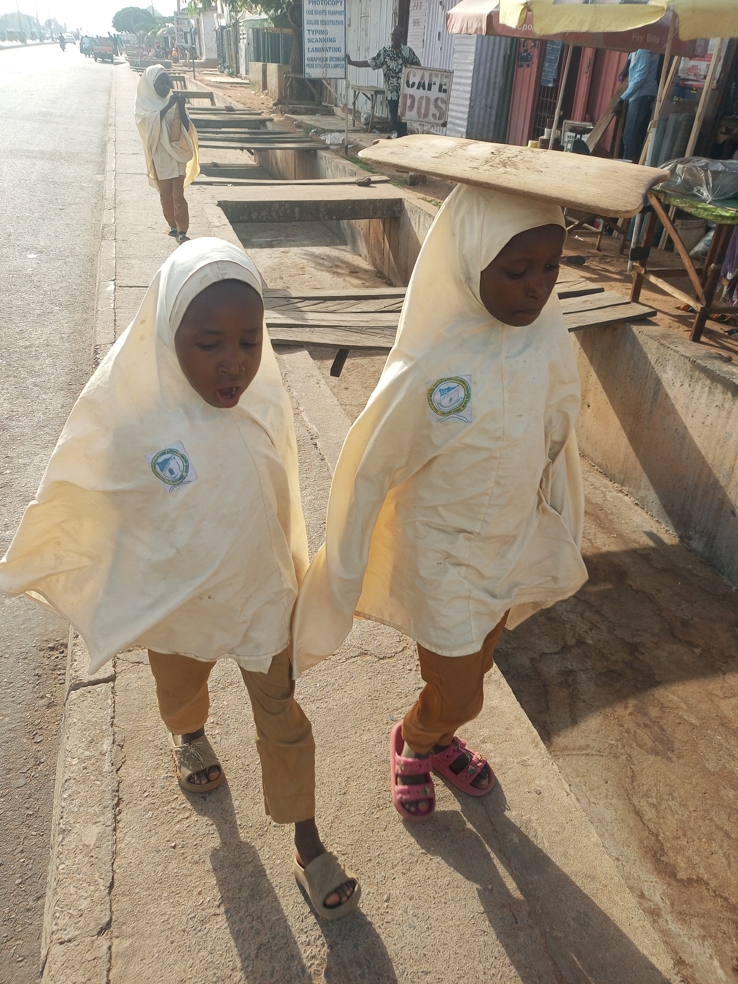 The image shows three young girls walking along a sidewalk in what appears to be Tempure, Nigeria. The girls are dressed in matching white headscarves and tunics with emblems on the chest and brown pants. The first two girls are in the foreground. The girl to the right has a large, flat board balanced on top of her head. They are walking next to an open drainage ditch along the sidewalk. In the background, there is another girl and a street scene with buildings and vehicles. There are signs advertising photocopying, scanning, graphic design, and cafe POS.