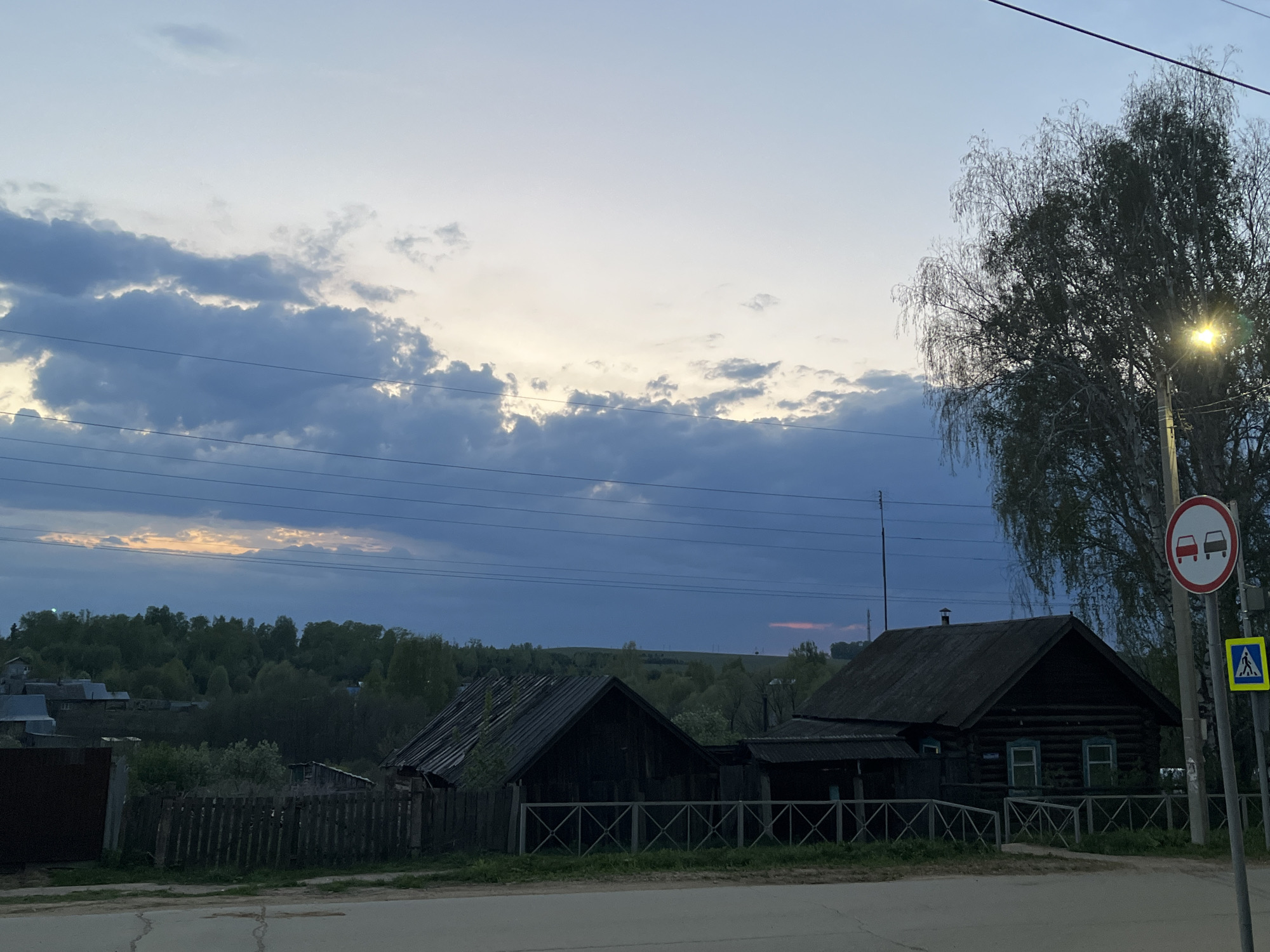 The scene depicts a rural area in Фоки, Russia, at dusk or dawn. In the foreground, a street with asphalt pavement runs horizontally. Along the roadside, there is a wooden fence and several old, dark wooden houses. A road sign indicating "No Overtaking" is visible next to a pedestrian crossing sign. A tree stands beside the sign, illuminated by a street light. The sky is filled with clouds, with faint orange light peeking through, suggesting sunrise or sunset. In the background, there is a distant treeline, suggesting a countryside environment. Overhead, several power lines stretch across the sky.