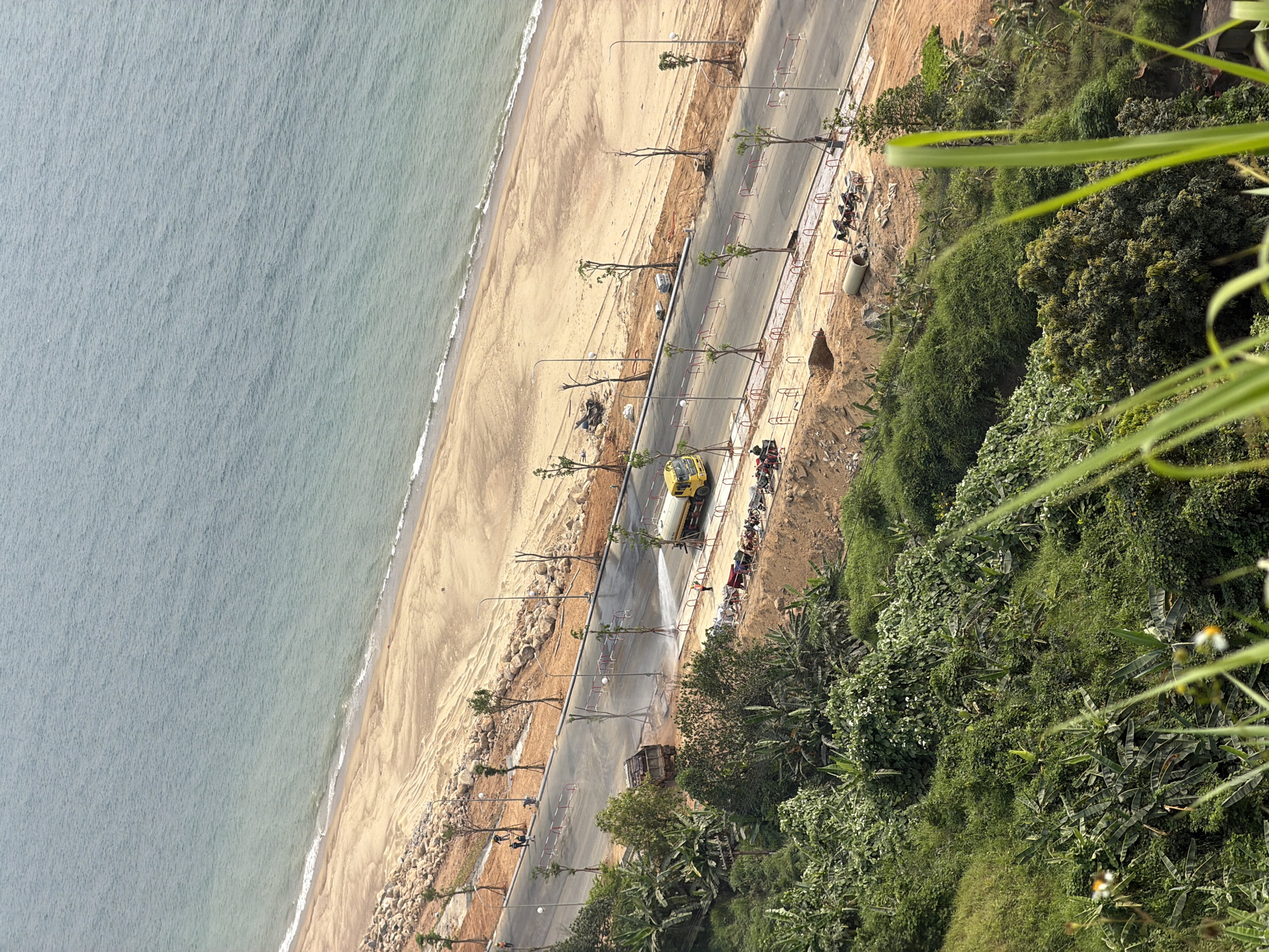 The image shows a coastal scene in Đà Nẵng, Vietnam. From an elevated vantage point, the view encompasses the beach, a road running parallel to the coast, and a dense, green hillside. The sea is on the left, with gentle waves meeting a sandy beach. Adjacent to the beach is a road with evenly spaced trees along its edge. A yellow vehicle is spraying water onto the road. There are parked motorcycles along the roadside, and a row of red and white barriers. On the right, the view extends to a lush, green hillside. The light is bright, suggesting a sunny day.