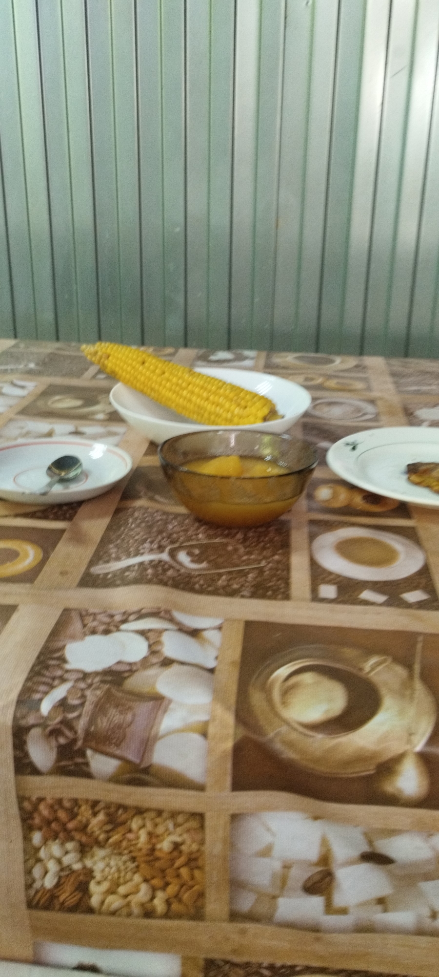 A close-up, slightly blurry, vertical shot showcases a table covered with a patterned cloth, featuring images of coffee beans, nuts, and possibly chocolate. On the table, a partially cooked ear of corn rests in a white bowl. To the right of the corn is a clear glass bowl filled with a yellowish liquid and pieces of fruit. Further to the right, a white plate holds a small piece of dark food, possibly a pastry or meat. To the left of the corn, another white plate contains a spoon. The background is a wall made of vertical metal panels, likely in a kitchen or dining area. The overall lighting is soft and even, suggesting an indoor setting. The image does not provide any clues about the location in Yaroslavskaya, Russia.