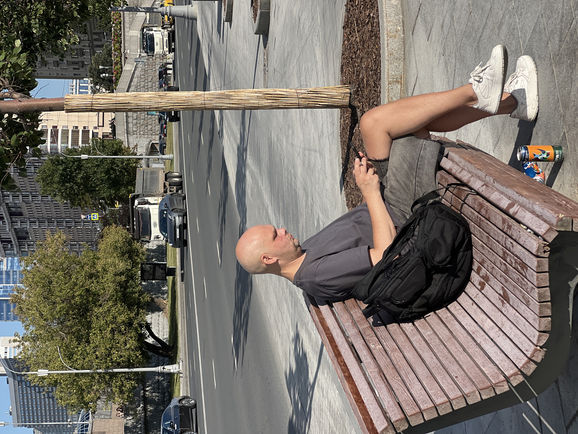 A bald, light-skinned man is sitting on a wooden park bench in Moscow, Russia, looking down at his phone. He is wearing a gray t-shirt and dark shorts, with white sneakers and socks. A black backpack is on the bench next to him. A can of beer is on the ground by his feet. In the background, traffic is moving on a multi-lane road, with cars, trucks, and a yellow taxi visible. Buildings and trees line the street, and a lamppost stands near the bench. The day appears sunny.
