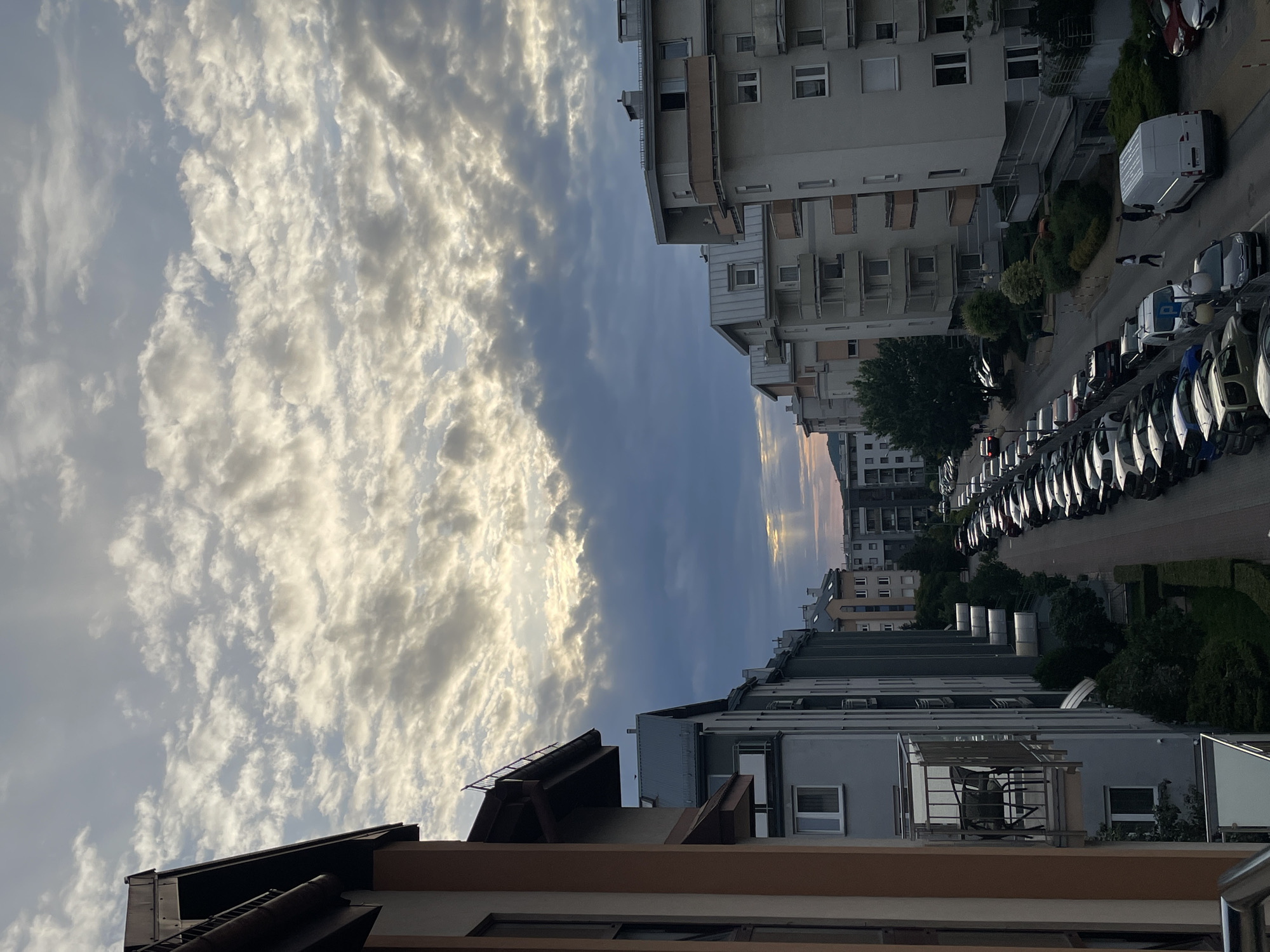 The image shows a street lined with apartment buildings in an urban setting. The sky is partly cloudy with rays of sunlight breaking through, suggesting it is either sunrise or sunset. Cars are parked along the street, and a few people are visible walking. The architecture of the buildings appears to be modern, with balconies and multiple stories. The overall scene depicts a typical residential street in a city.
