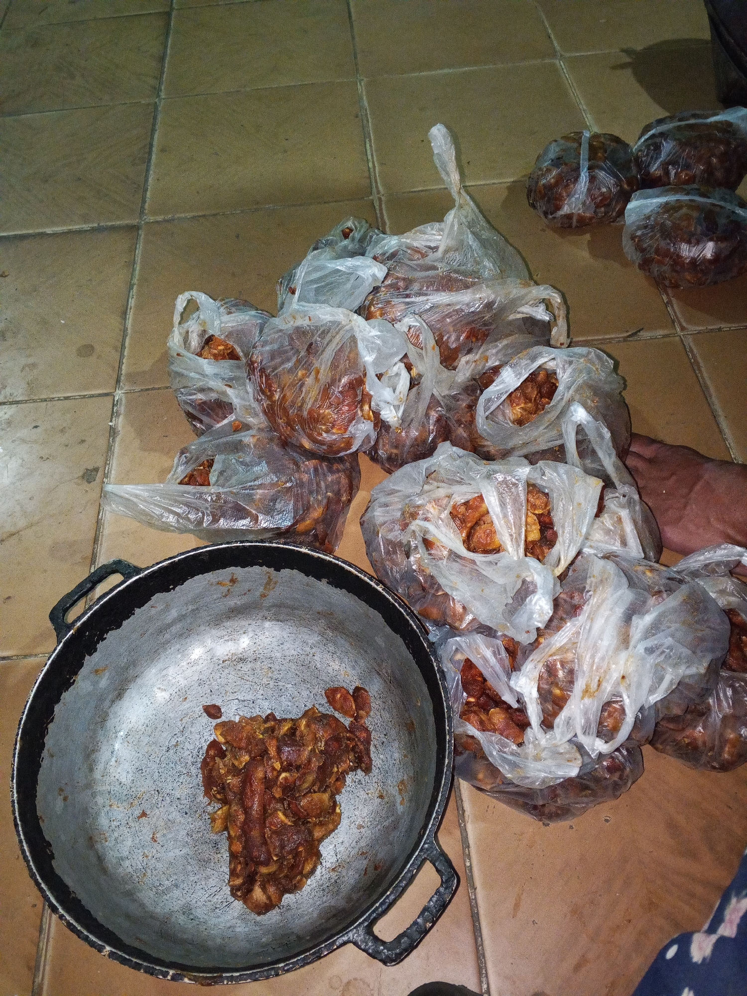 The image shows a pile of tamarind fruit packaged in clear plastic bags. A large metal pot sits at the bottom left corner of the image, containing a portion of tamarind. The bags are arranged on a tiled floor. The image was taken indoors. There is a foot visible in the lower right portion of the image. The location is Altagracia, Venezuela.