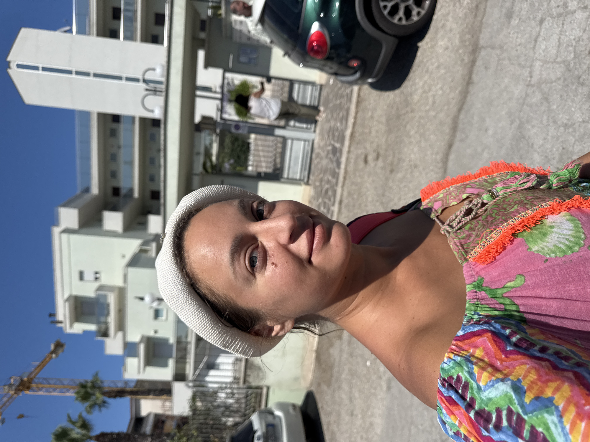 A woman wearing a white hat and an off-the-shoulder, colorful dress is taking a selfie in front of a modern building. The building appears to be a hotel or apartment complex with balconies and large windows. A yellow construction crane is visible in the background, along with palm trees. A green car is parked on the right side of the image, and a white car is parked on the left. The scene is outdoors on a sunny day. The woman's dress has a vibrant pattern with shades of pink, orange, yellow, blue, and green. It is embellished with orange fringe and braided cords with tassels. The woman has light skin, blue eyes, and her hair is pulled back. The environment suggests a coastal city, possibly San Benedetto del Tronto, Italy, given the presence of palm trees and modern architecture.