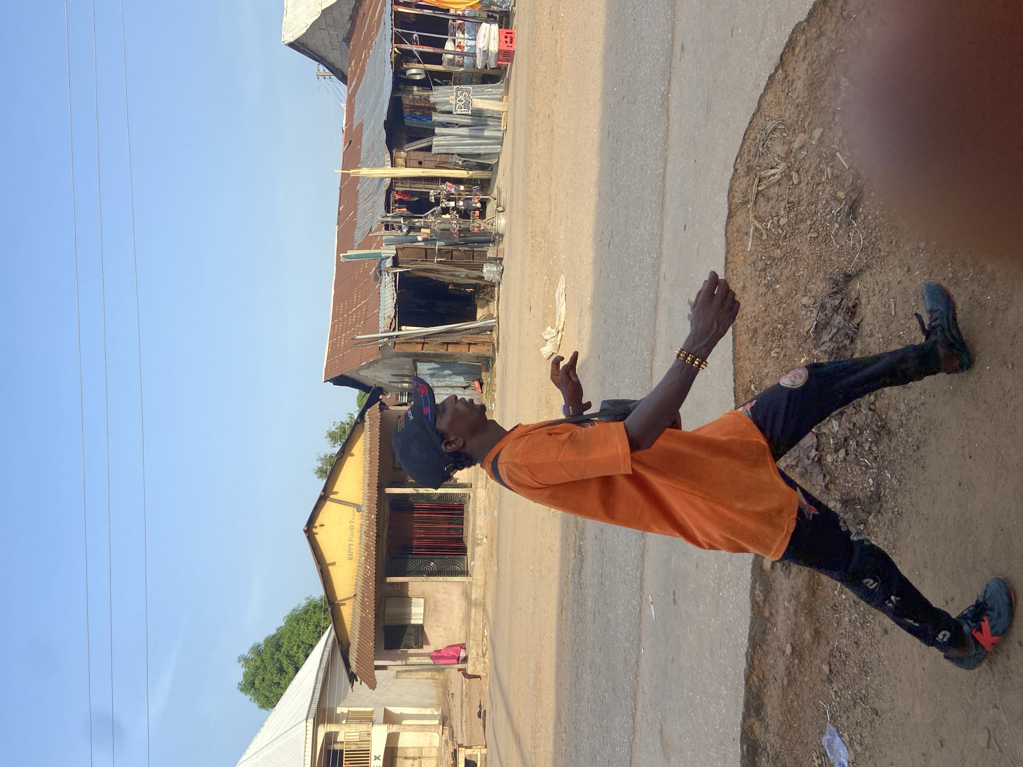 In Mararaba, Nigeria, a slender black man, facing left, wears an orange t-shirt, black leggings, and dark sneakers. He is also wearing a dark baseball cap. The man stands on a pavement's edge where it transitions to dirt. Behind him, buildings vary in construction and age, with corrugated metal roofs. Overhead, wires stretch across the sky. The photograph is taken in daylight.