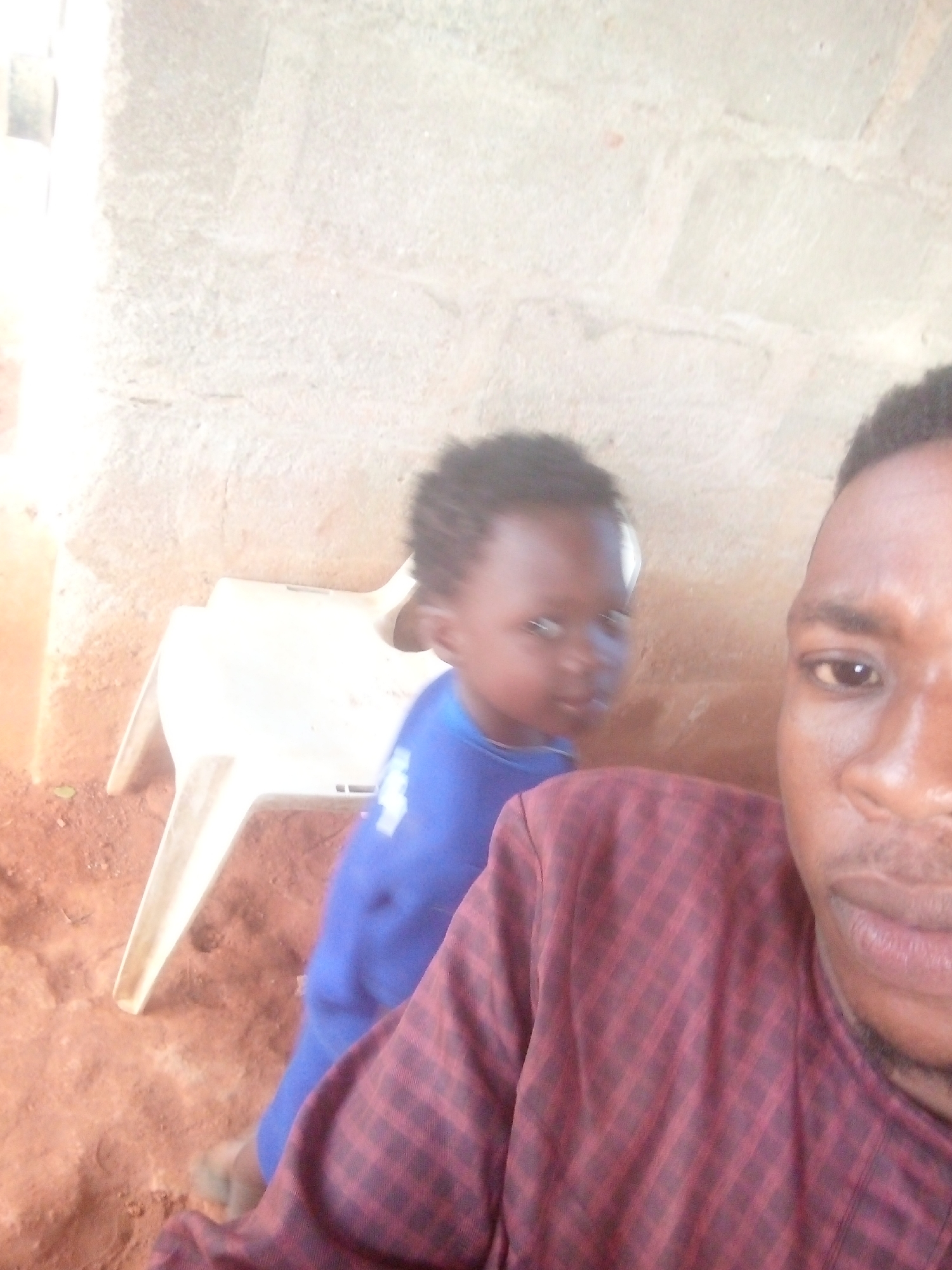 A man and a child are located in an outdoor environment in the city of Unknown, Unknown. The man is partially visible in the foreground, wearing a maroon plaid shirt. The child stands behind a white plastic chair, wearing a blue shirt. The backdrop consists of a textured beige wall. The ground appears to be soil.