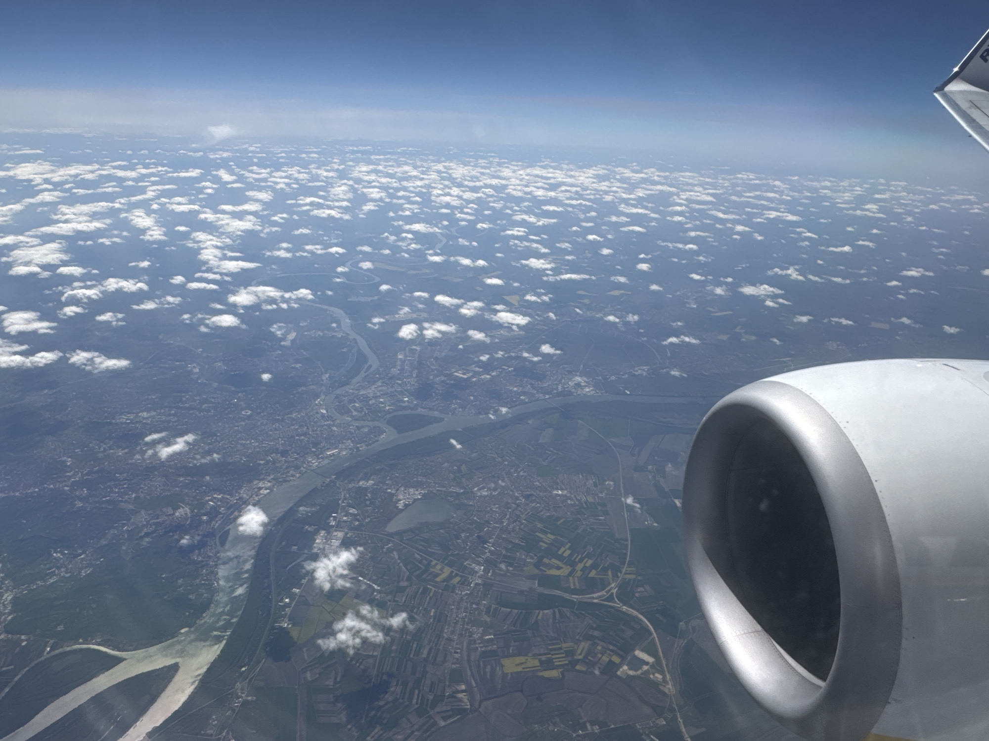 The image captures an aerial view from an airplane window, looking down upon a vast landscape under a clear blue sky dotted with scattered cumulus clouds. In the foreground, the silver nacelle of the aircraft's engine is prominent on the bottom right, with a small section of the wing visible at the top right, bearing the partial text "RY."
Below the scattered clouds, a significant river system winds through the terrain. A wide, main river is joined by a substantial tributary in the mid-ground, creating a prominent confluence. Extensive urban areas are built along the riverbanks, characterized by dense clusters of buildings and visible infrastructure like roads and what appear to be bridges. Beyond the urban sprawl, the landscape transforms into a patchwork of agricultural fields, displaying varied shades of green and yellow, interspersed with areas of denser forest or woodland.
The scene is brightly lit, indicating daytime, likely mid-morning to early afternoon, under fair weather conditions with excellent visibility. While the provided location context is Sofia, Bulgaria, the depicted geographical features, specifically the large river confluence and the scale of the associated major urban development, do not align with the geography of Sofia, which is an inland city. The visual evidence strongly resembles the confluence of the Danube and Sava rivers with the city of Belgrade, Serbia, sprawling along their banks.