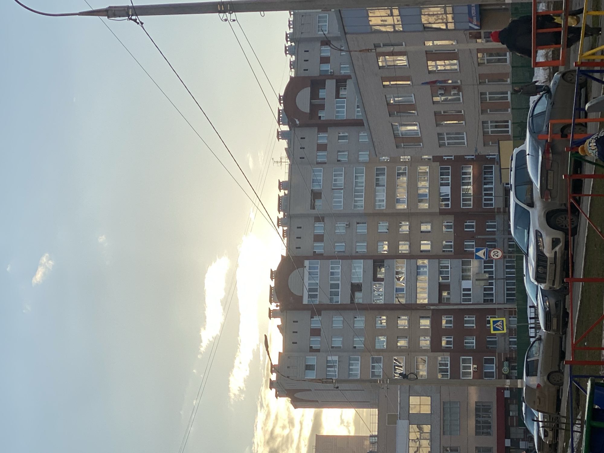 The image shows a residential area in Izhevsk, Russia, with a large apartment building dominating the scene. The building has a distinctive architectural design, featuring a rounded top section. The sky is bright, with the sun partially obscured by clouds, suggesting either sunrise or sunset. Power lines run across the sky, adding to the urban setting.
Several cars are parked along the side of the building. Part of a playground is visible on the right. There is a pedestrian crossing street sign on the right. A person wearing a red hat is standing on the playground. A Russian flag can be seen on the building.