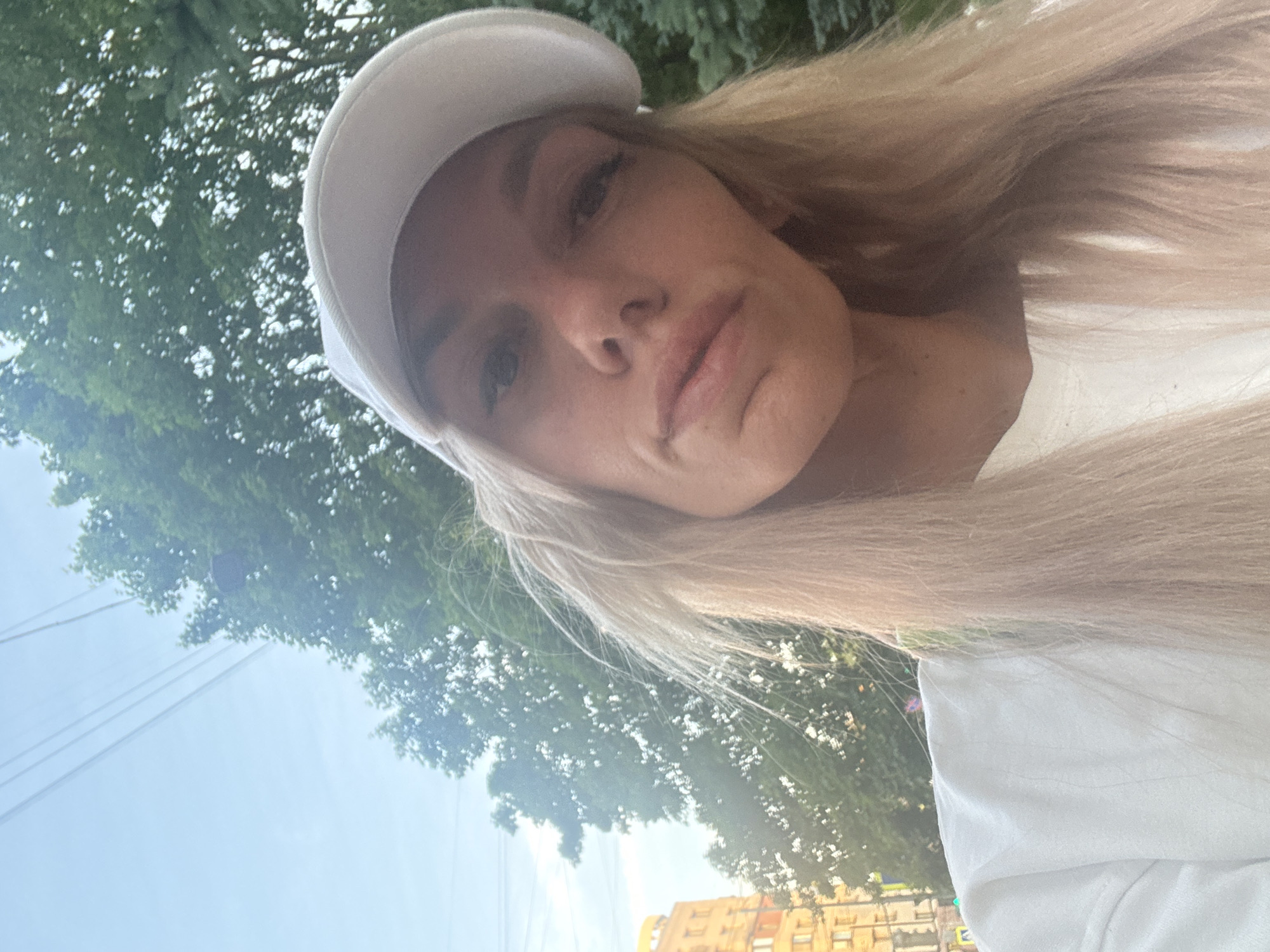 A woman wearing a white cap and a white sweatshirt is taking a selfie outdoors. The background shows trees, a building, and power lines, suggesting an urban environment, possibly in Saint Petersburg, Russia, given the architectural style of the building. The woman has blonde hair and is looking at the camera. The lighting suggests it is daytime.