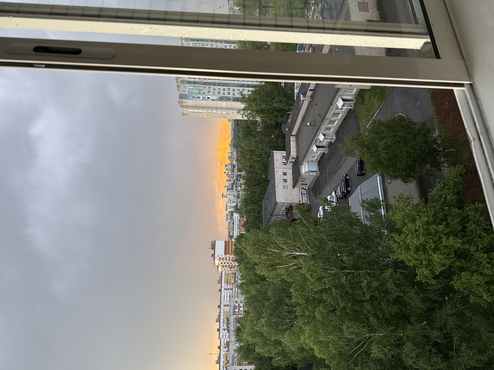 The image shows a view from a high-rise building in Izhevsk, Russia, looking out over the city. A partially open window frame is visible at the top and sides of the frame. The sky is predominantly gray and overcast. An orange hue is visible on the horizon where the sky meets the buildings. Below the horizon, multiple tall buildings can be seen intermixed with trees and other vegetation. The photograph also includes a small section of an apartment courtyard. A group of vehicles is parked in a small parking lot near a grey building. The foreground is primarily made up of green tree canopies.