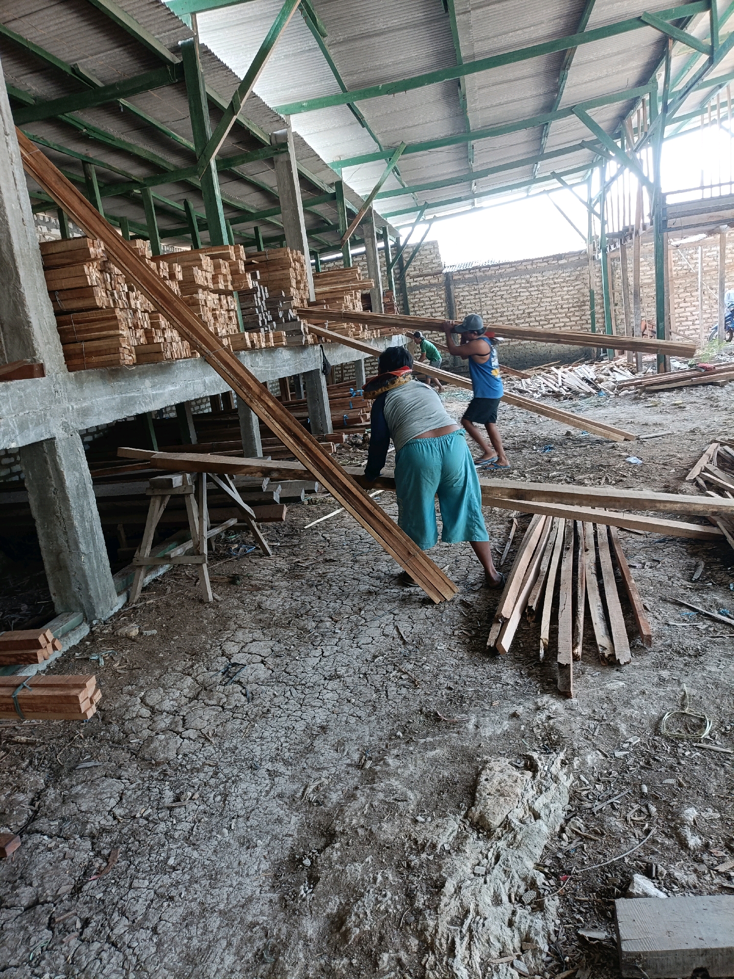 The image depicts an outdoor lumberyard or workshop in Pordapor, Indonesia. Two men are actively working with lumber. In the foreground, a man wearing blue shorts and a grey long-sleeved shirt is carrying a piece of lumber on his shoulder. Behind him, another man in a blue tank top and a grey cap is also carrying lumber. They are surrounded by stacks of wood, building materials, and the interior of a structure with a corrugated metal roof supported by green metal beams. The ground is uneven and appears to be dry, cracked earth mixed with debris. In the background, a brick wall and more lumber are visible. The overall scene suggests a busy working environment for lumber processing or construction.