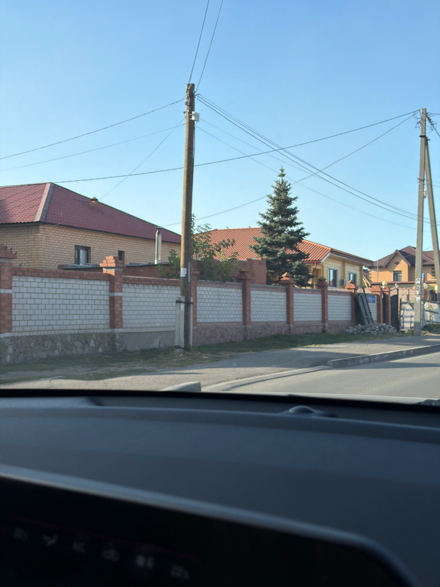 The image shows a street in Pavlodar, Kazakhstan, with a brick fence lining the property. The fence is made of white brick with red brick pillars and accents. Behind the fence, there are houses with red and yellow roofs, and a tall evergreen tree. Power lines are visible in the sky, supported by wooden and concrete poles. The foreground shows the dashboard of a car. The sky is clear and blue. The image appears to be taken during daylight.