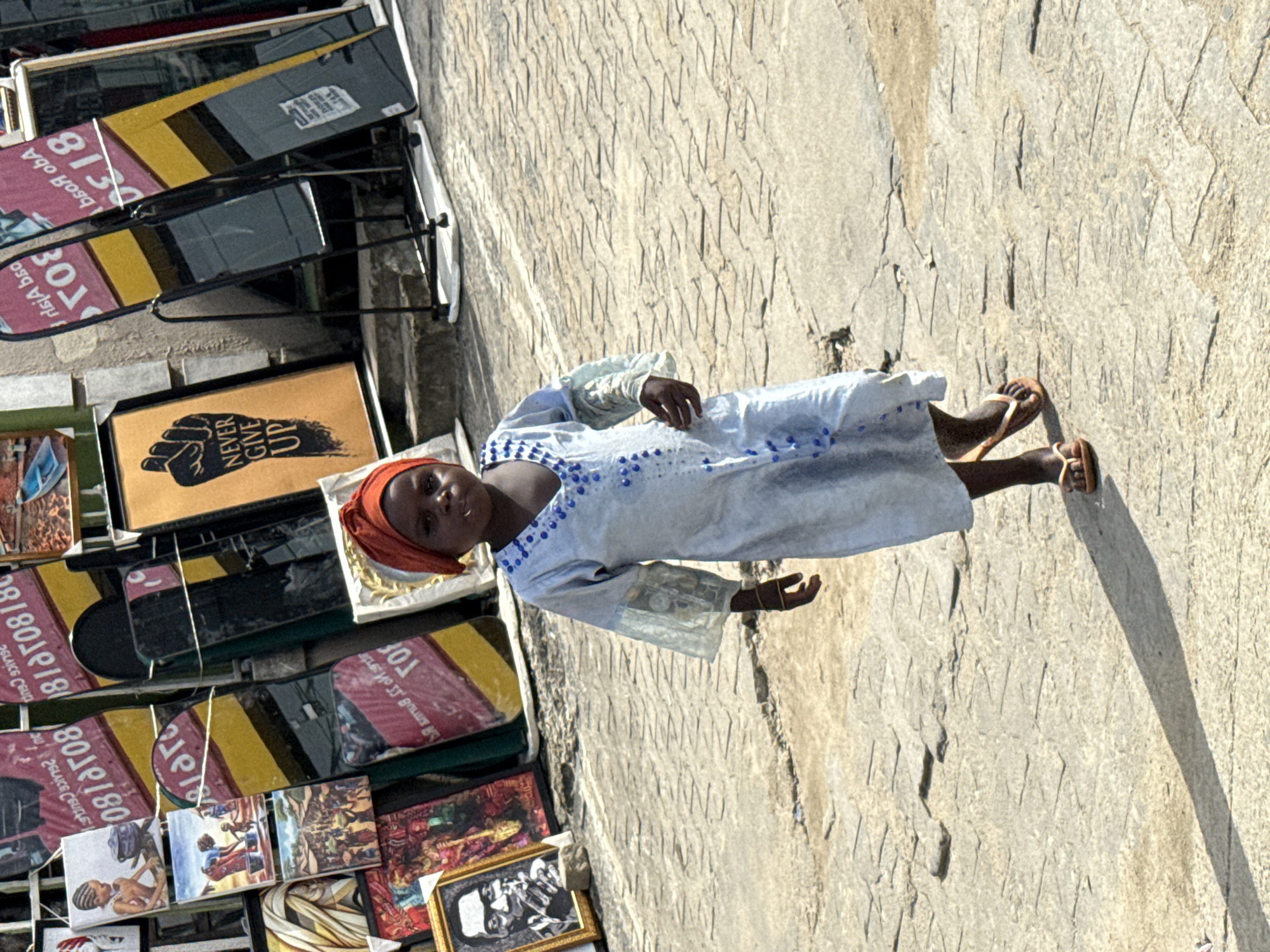A full shot, taken during daylight hours, depicts a young dark-skinned girl in Ado, Nigeria, standing on a concrete surface, possibly a sidewalk or street edge. She is clad in a light-colored, long-sleeved dress with blue embellishments, a red headwrap, and sandals. Her direct gaze towards the camera suggests a sense of awareness or interaction with the photographer.
To the left of the girl, an assortment of framed prints and artwork is displayed on a portable rack. These prints feature various subjects, including portraits, graphic designs ("Never Give Up" is visible), and commercial branding. The rack itself appears to be a simple metal structure designed for outdoor display.
The background consists of the concrete surface and the shadows cast by the girl and the rack, indicating the presence of a light source, likely the sun. The environment suggests an urban or commercial area, with the display of artwork implying a market or business setting. The composition is oriented vertically, with the girl positioned slightly off-center.