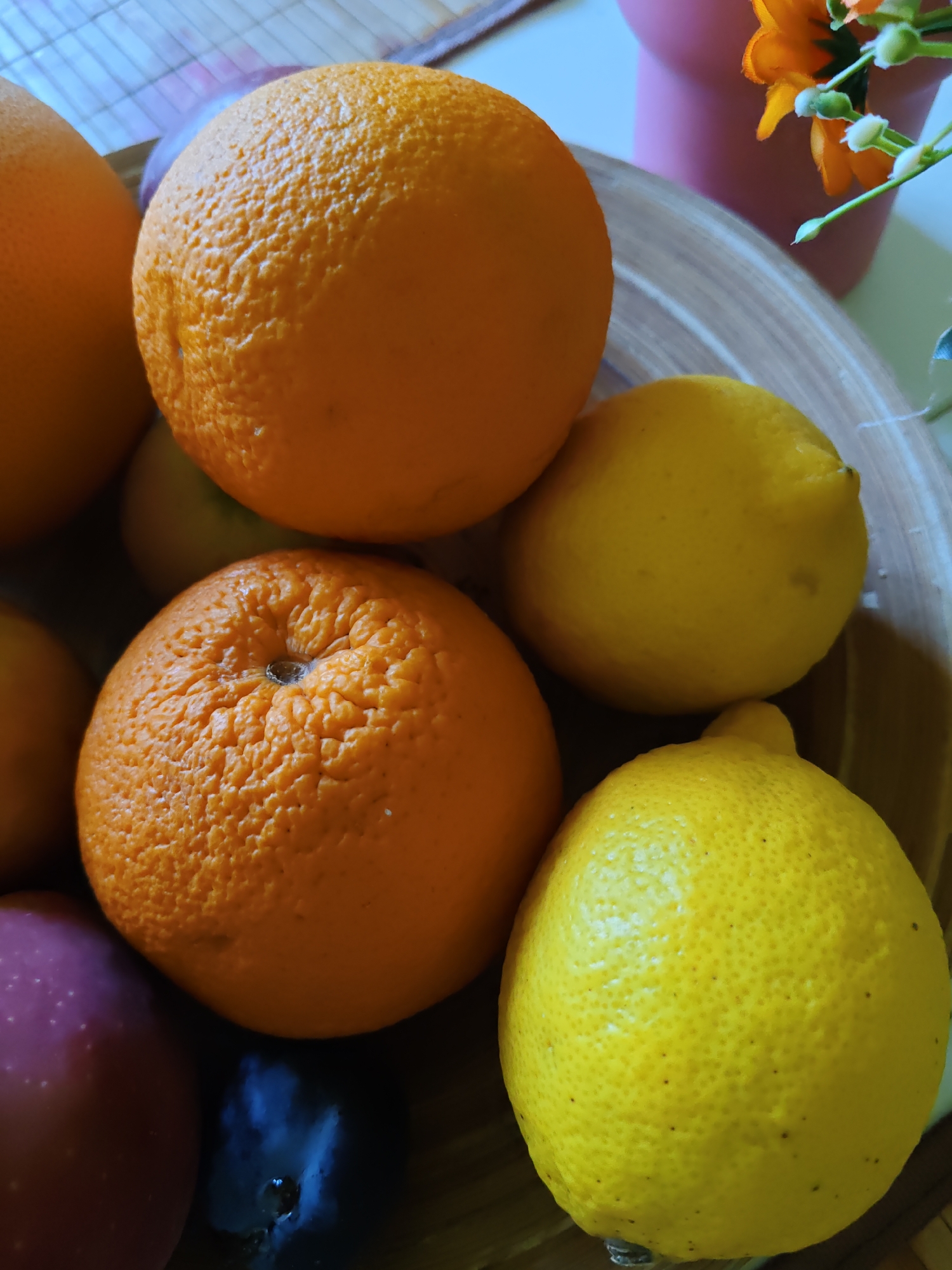 The image shows a close-up, overhead view of a collection of fruits in a wooden bowl. The bowl contains two oranges, two lemons, and a dark-colored fruit, possibly a plum, and a red apple. The fruits are whole and appear fresh. In the background, a pink vase with orange flowers is visible, as well as part of a table with a patterned surface. The lighting suggests an indoor setting, possibly a kitchen or dining area. The image does not contain any information related to Rostov-on-Don, Russia.