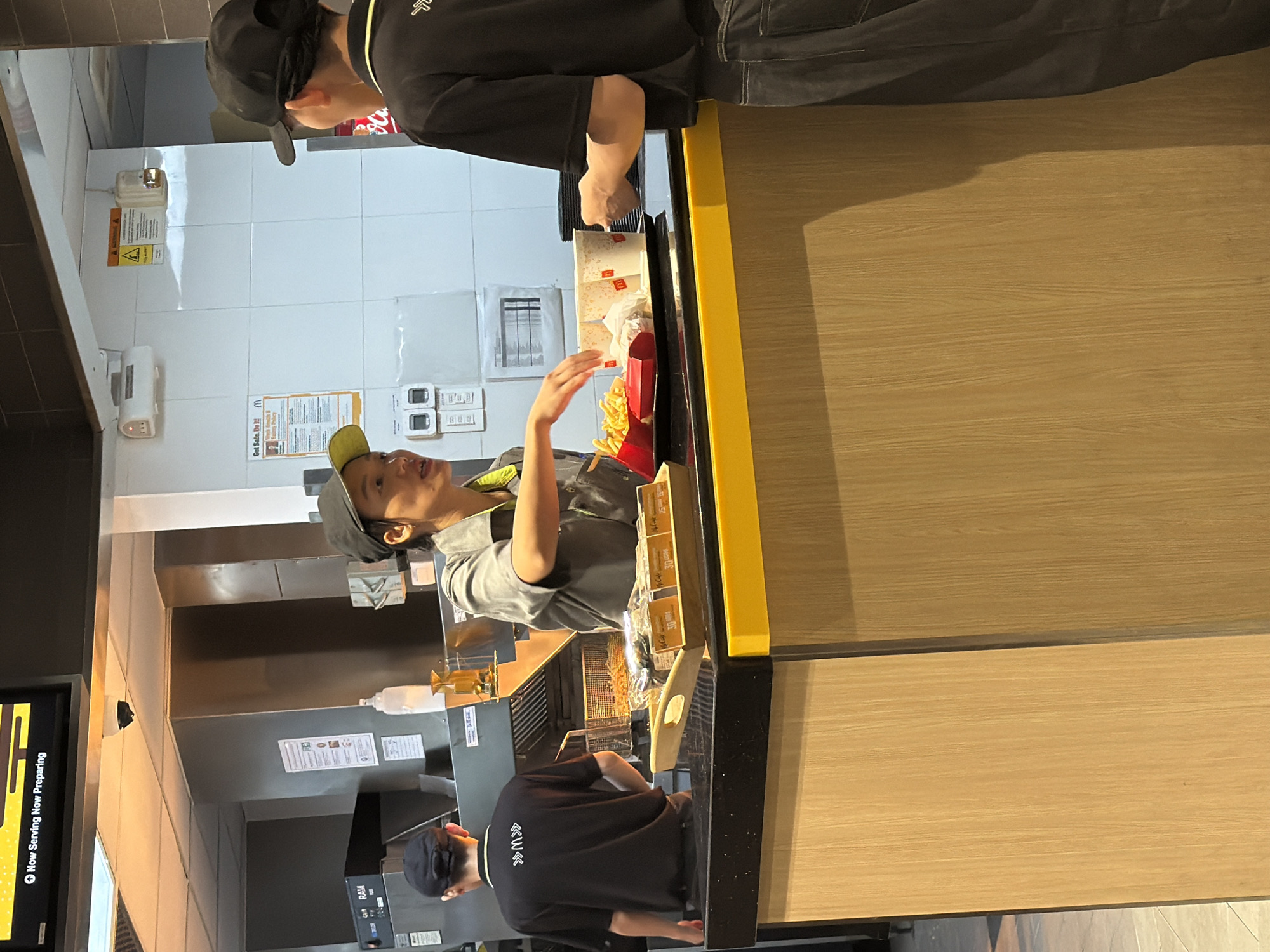 In a fast-food restaurant, possibly McDonald's, in Nha Trang Ward, Vietnam, two individuals are engaged in a transaction. On the left, a young person wearing a black cap and a black t-shirt with white insignia on the back is standing and looking towards a counter. On the right, another individual, also wearing a black cap and a black t-shirt, is facing away from the camera, leaning over the counter. Between them, a female employee in a grey uniform with a yellow collar is interacting with the customer, handing them food and drinks. A tray on the counter holds red containers filled with french fries and two McDonald's branded cups. The background reveals kitchen equipment, including a deep fryer, and tiled walls. The counter area is equipped with a yellow trim along the top. A digital display above the counter shows "Now Serving Now Preparing".