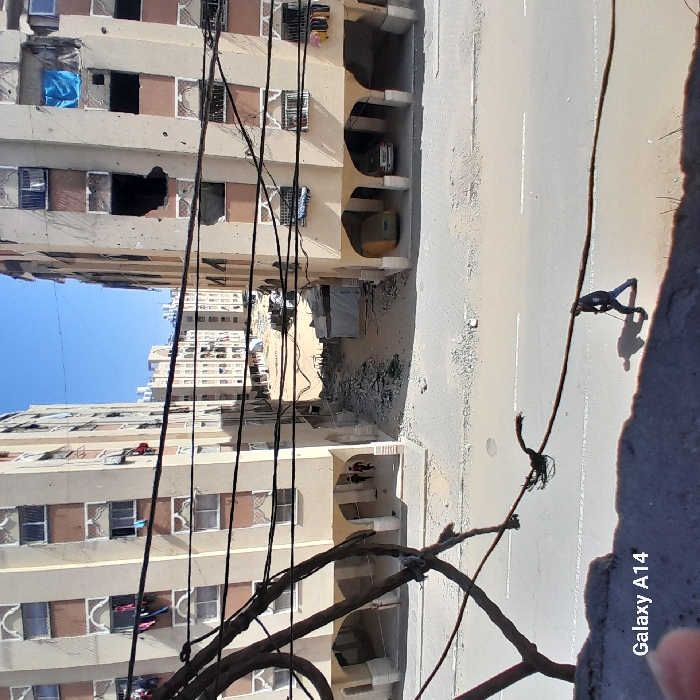 The image depicts a damaged urban residential environment in Khan Yunis, Palestinian Territory, viewed from an elevated position. Two multi-story apartment blocks with beige and brown facades dominate the scene, flanking a central, rubble-strewn expanse. Both buildings exhibit extensive damage, including numerous perforations consistent with projectile impacts across their surfaces and large, irregular apertures on upper floors, particularly prominent on the right-hand building. Some windows are missing panes, exposing dark interiors, while others show clothing hanging outside.
Between the two primary buildings, the ground is an unpaved area filled with construction debris, concrete fragments, and scattered rubble, suggesting demolition or significant structural damage. Further back, additional damaged apartment blocks are visible under a clear blue sky, interspersed with what appear to be makeshift shelters or structures within the debris field.
In the immediate foreground, a concrete barrier or ledge partially frames the lower edge of the image, with multiple dark utility cables crossing horizontally. Below these cables, a paved street runs left to right. A lone individual, dressed in dark clothing, walks on the street from right to left, leaning on a stick or cane and casting a distinct shadow. Under the arched ground-floor openings of the left building, two smaller figures are seen walking away from the camera. The ground floor arches of the right building contain a yellow van and a silver sedan. The watermark "Galaxy A14" is present in the lower-left corner of the image.