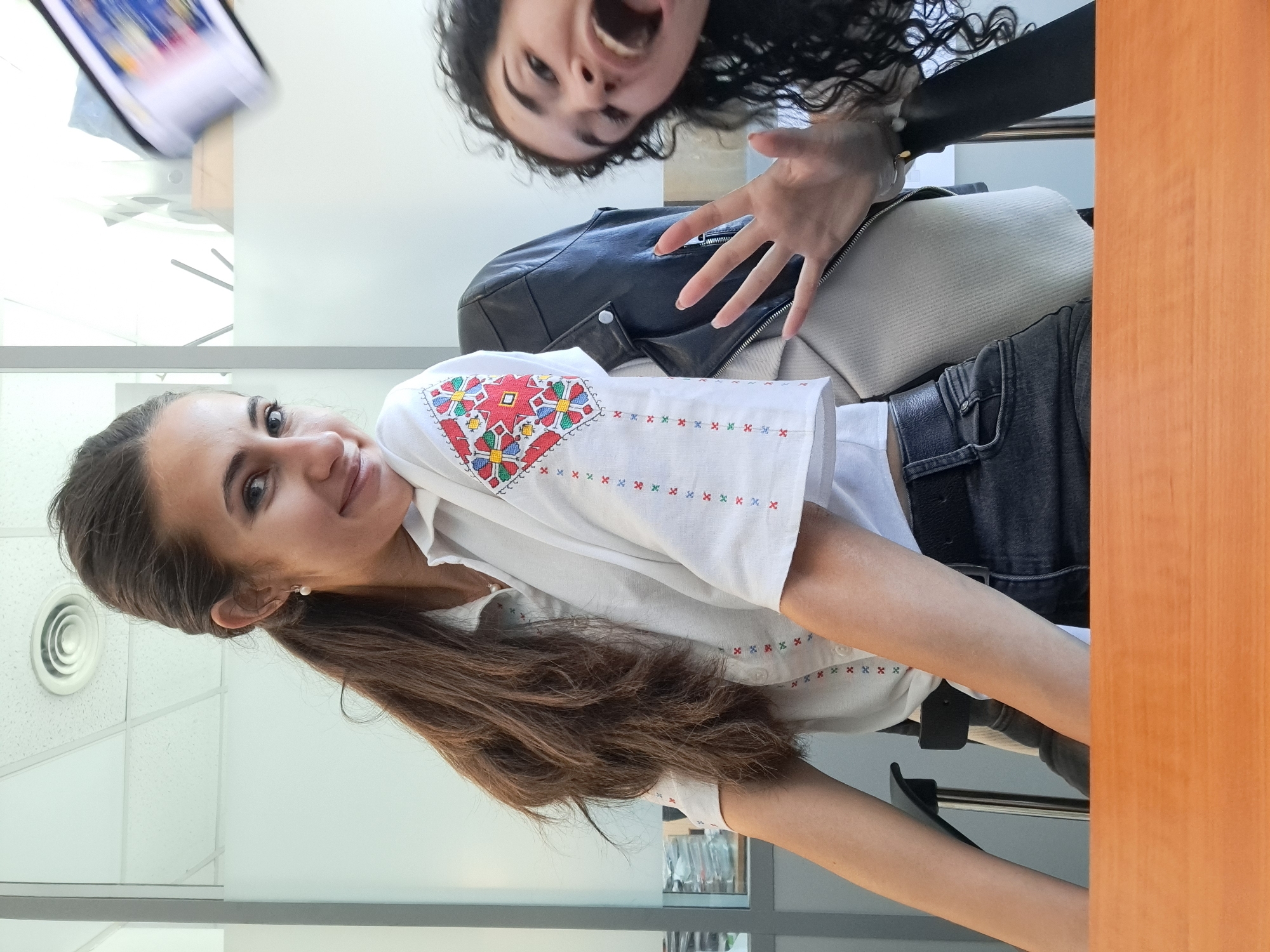 Two young women are featured in a bright, indoor setting, likely captured in a playful selfie. The image is rotated 90 degrees counter-clockwise, making the subjects appear tilted.
In the foreground, a woman with long, dark brown hair smiles gently towards the camera. She wears a white shirt with distinctive, colorful cross-stitch embroidery. The embroidery on her right sleeve displays red, blue, green, and yellow geometric patterns, while smaller 'X' motifs in similar colors decorate the rest of the shirt. A small pearl earring is visible on her ear.
Behind her, appearing inverted relative to the frame (suggesting she is closer to the camera and looking down into it), is another woman with dark, curly hair. She has a wide-open mouth, conveying an expressive, playful, or excited emotion, and her right hand is raised. She is wearing a black leather jacket over a light-colored top and dark jeans. A beige bag or cushion is partially visible beneath her jacket.
The background reveals a modern indoor environment, possibly an office or classroom. Features include white ceiling tiles, a circular air vent, and light-colored walls. A blurry screen (likely a tablet or smartphone) showing indistinct content is visible in the upper left corner. On the right side of the frame, a warm-toned wooden surface, possibly a desk or partition, is prominent. Parts of a black chair and dark-colored jeans are also seen.
The scene conveys a cheerful and casual interaction between the two individuals. The lighting is bright, indicating daytime. No discernible text is visible on any objects or in the background. While the location context is Sofia, Bulgaria, there are no specific visual cues within the image to confirm this.