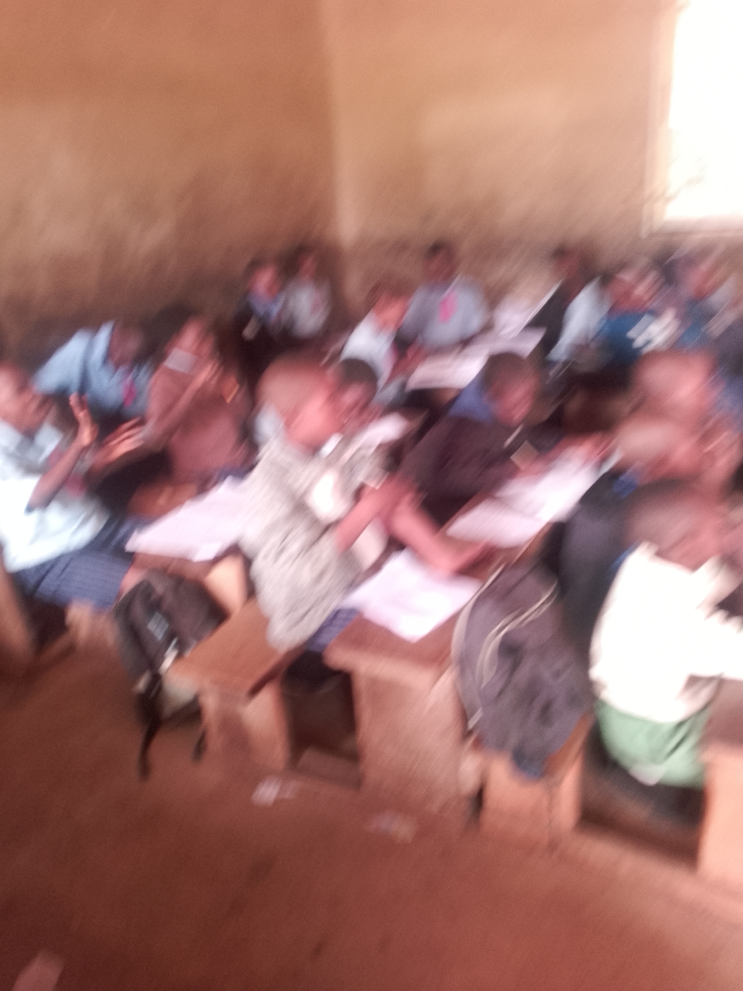 A group of people, likely students, are seated at wooden desks in a classroom. The image is blurry, making it difficult to discern specific details, but it appears to be an indoor setting with plain walls and a window with natural light. Some individuals are wearing shirts and ties, suggesting a formal or school environment. Papers are spread across the desks, and some people are looking towards the front of the room, possibly at a teacher or a presentation. Backpacks and other personal belongings are visible near the desks. The overall impression is one of a classroom lesson or activity in progress. The location is identified as Unknown, Unknown.
