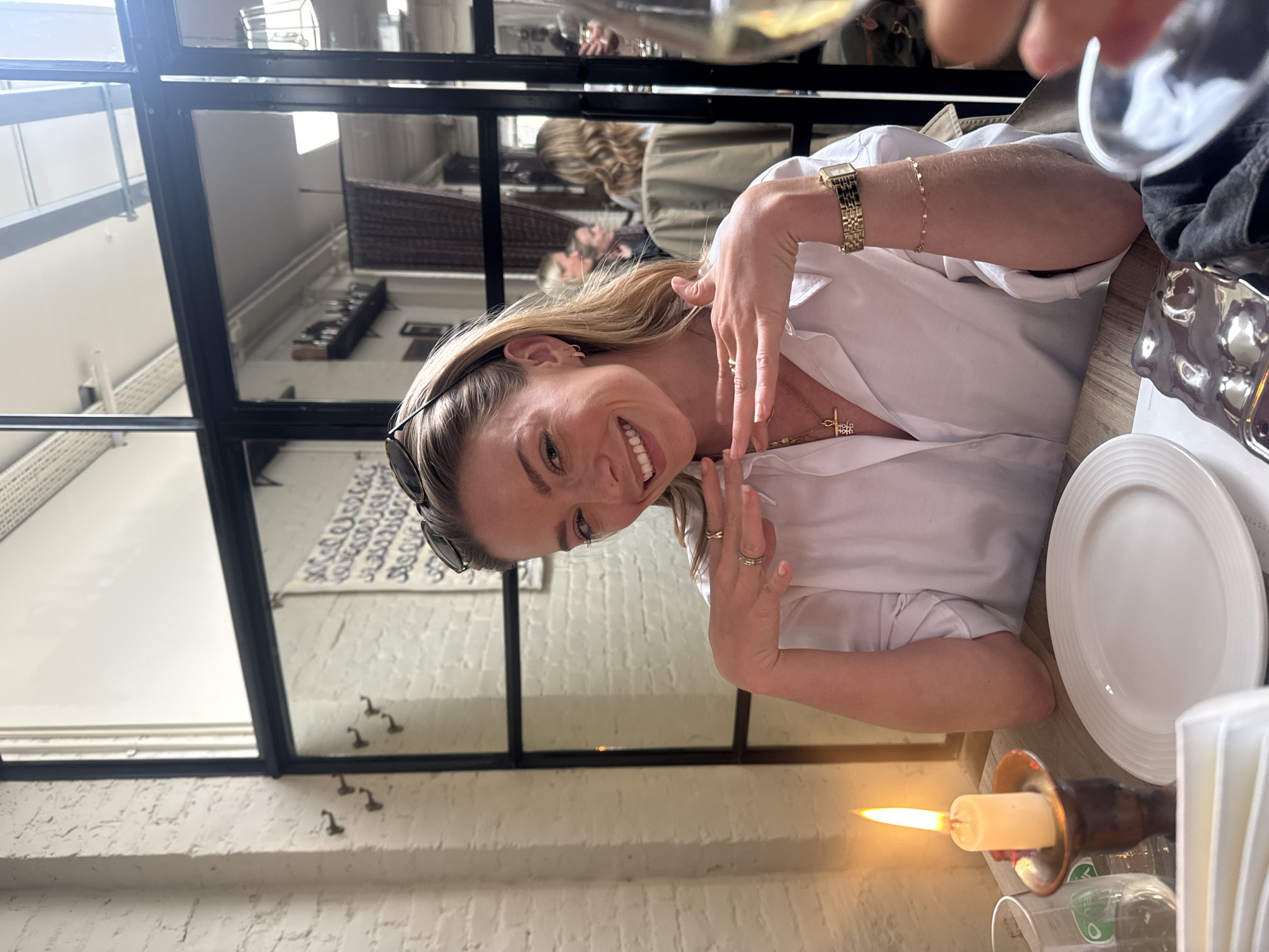 The image depicts a woman sitting at a table in a restaurant. She is wearing a white shirt and gold jewelry, including a watch, bracelets, and necklaces. Her hands are raised near her face. On the table, there is a white plate, a decorative metal object, a candle holder with a lit candle, and a wine glass. A window with black frames is visible behind her, reflecting the interior of the restaurant. The location is in the city of Unknown, Unknown.