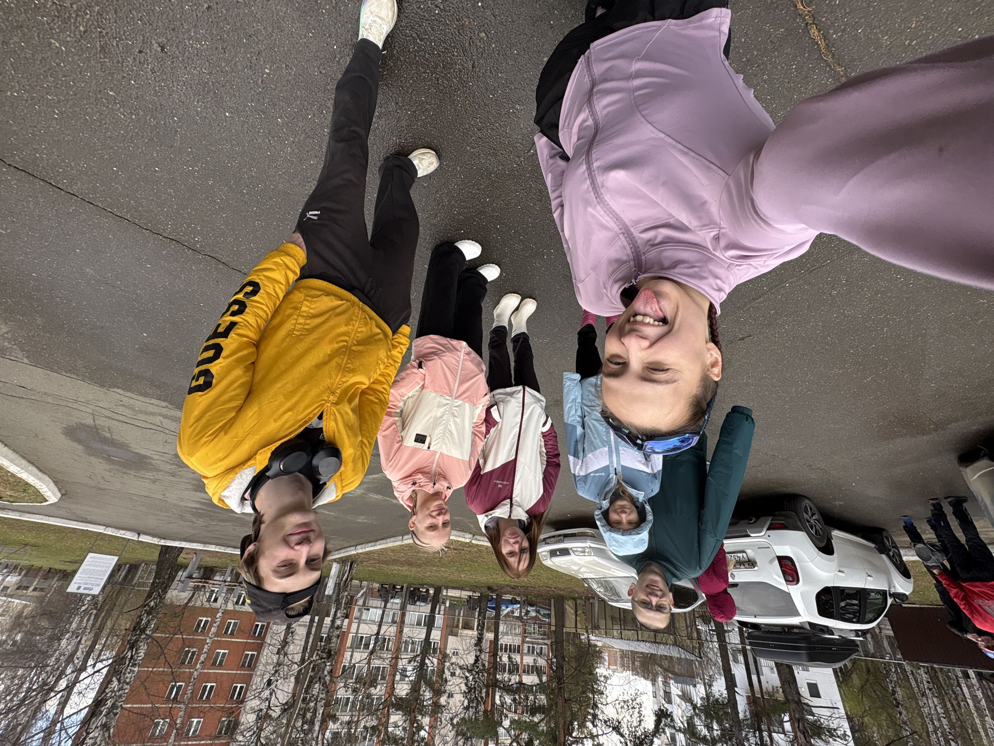 Six people are posing for a selfie taken from a low angle on a paved surface, likely a street or parking lot in Chaikovsky, Russia. The buildings in the background appear to be residential.
The person taking the photo is in the upper right and is wearing a light purple jacket and has her tongue out. To her left and slightly behind is a person in a blue jacket and glasses. To their left, a person is wearing a white and red coat. To their left a person is wearing a pink and white coat. To their left a person is wearing black pants and a yellow coat. To their left is a person in black pants and a white shoe.
Behind the people, a white car with a roof cargo carrier is parked. Trees are visible, with some appearing bare, indicating it may be either early spring or late autumn. A sign and a building are visible in the background. Some people are in the background, to the right of the car.