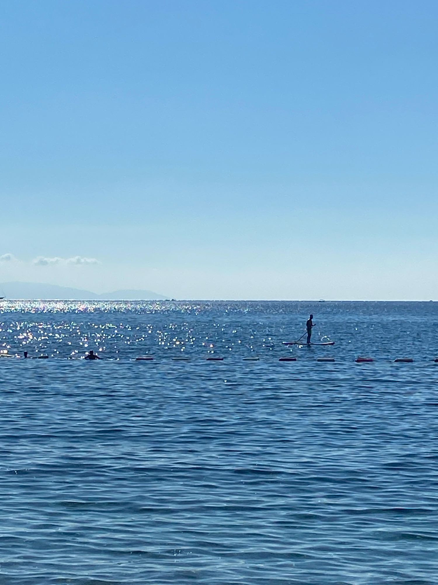 A silhouetted person stands on a paddleboard in the middle of a calm, blue sea. The sun glints off the water's surface, creating a shimmering effect. In the distance, hazy mountains are visible under a clear, pale blue sky. A rope line with buoys runs horizontally across the water, separating the foreground from the middle ground. A few other swimmers are visible in the water closer to shore. The scene appears to be a serene beach or coastal area.