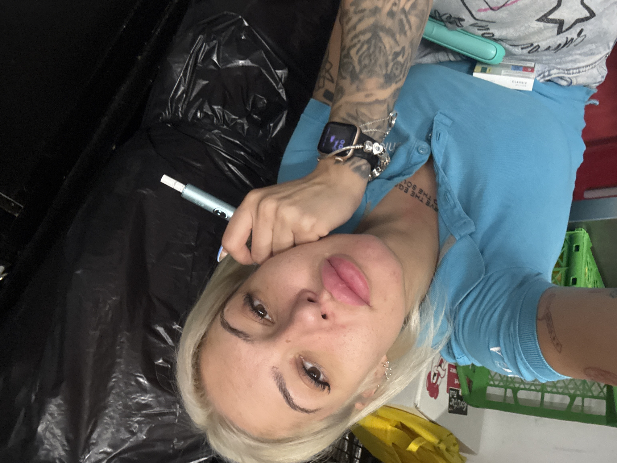 A female individual, appearing to be an adult with blonde hair, defined dark eyebrows, pronounced lip fillers, and eyelash extensions, is depicted in a medium close-up, gazing directly at the viewer. She wears a light blue polo shirt with an open collar and light-colored bottoms adorned with star patterns. Visible tattoos include partial text on her neck/upper chest (e.g., "RVE THE EG" and "THE SOU"), a full sleeve tattoo on her left forearm, and a smaller tattoo on her right bicep. Her left wrist displays a black Apple Watch with a rose gold-colored case, showing purple graphics, alongside multiple silver chain bracelets and charms. She holds a light blue and silver pen-shaped device, consistent with an IQOS heated tobacco holder, in her right hand, which also supports her head. Adjacent to her, a white and red box labeled "CLASSIC," likely containing HEETS tobacco sticks, and a teal-colored elongated object, possibly an IQOS pocket charger, are present. The background, an indoor environment in Plovdiv, Bulgaria, includes a green plastic crate, a yellow object, a white wall with a partially visible red and white chicken logo sticker, and dark, crumpled plastic sheeting. The scene is lit by overhead artificial lighting.