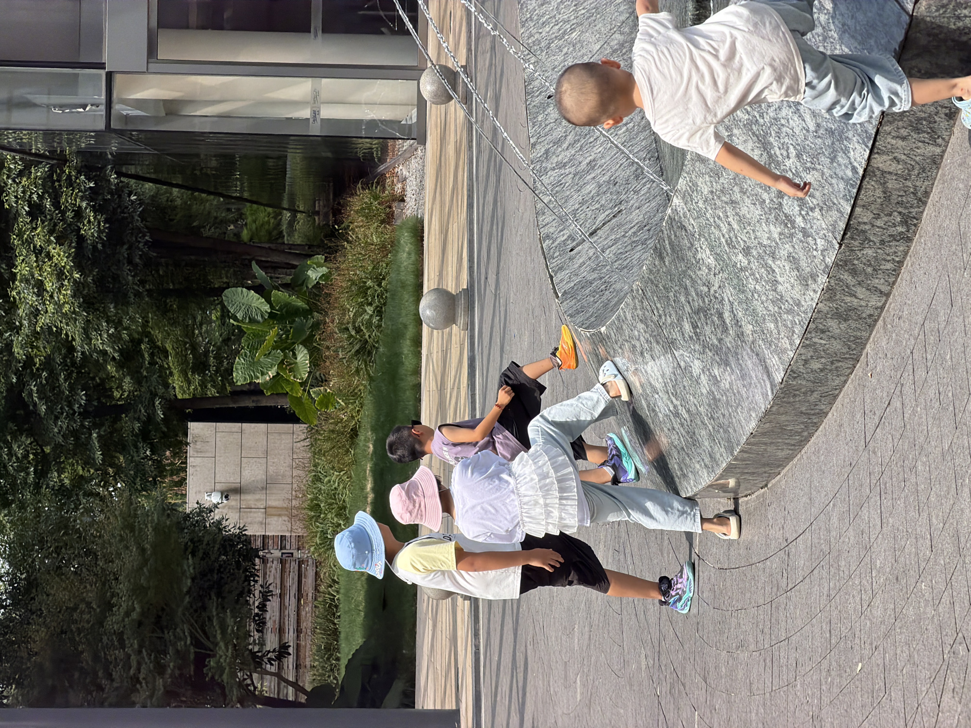 In the Yuzhong District, China, a sunny outdoor scene unfolds with a group of four young children near a water feature. Three children, facing away from the camera, are clustered together on a gray, textured surface with a curved edge. The leftmost child wears a yellow and white short-sleeved shirt, black shorts, and a light blue bucket hat. Next to them, a child in a white ruffled top, light blue pants, and a pink bucket hat appears to be looking down at the water. The third child, in a dark purple sleeveless shirt and black shorts, is seated on the edge of the water feature, wearing orange athletic shoes. A fourth child, facing away and slightly to the right of the others, wears a white t-shirt and light blue baggy shorts, with their back to the viewer. Water streams arc over the gray stone structure. In the background, lush green trees and foliage are visible, along with a light-colored brick wall and a modern building with glass windows. The ground is paved with gray square tiles arranged in a circular pattern.
