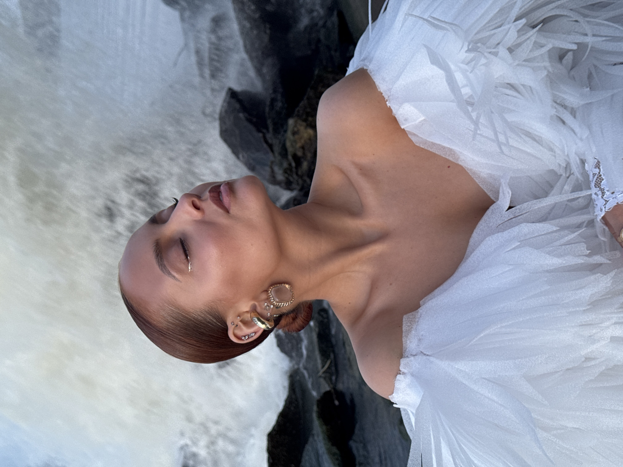 A woman with reddish-brown hair, fair skin, and closed eyes is posing in front of a waterfall near some rocks. She is wearing a white, feathered dress and gold earrings. The location may be in Chaikovsky, Russia, as indicated by the request.