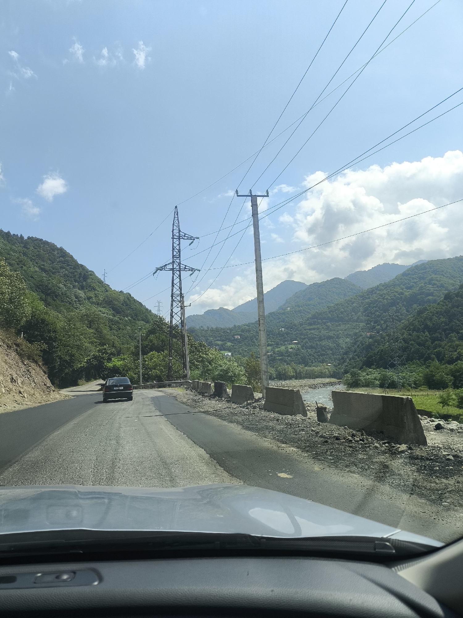 The image depicts a road with a car driving on it, heading towards a bridge with concrete barriers on the side. The road is surrounded by lush green mountains, and a river flows beside it. Overhead, a clear blue sky with scattered clouds is visible, along with power lines supported by tall metal towers. In the distance, more mountains and a small cluster of houses can be seen nestled amongst the trees, suggesting a rural setting within the city of Kveda Bzubzu, Georgia. The overall scene is one of a scenic drive through a mountainous landscape.