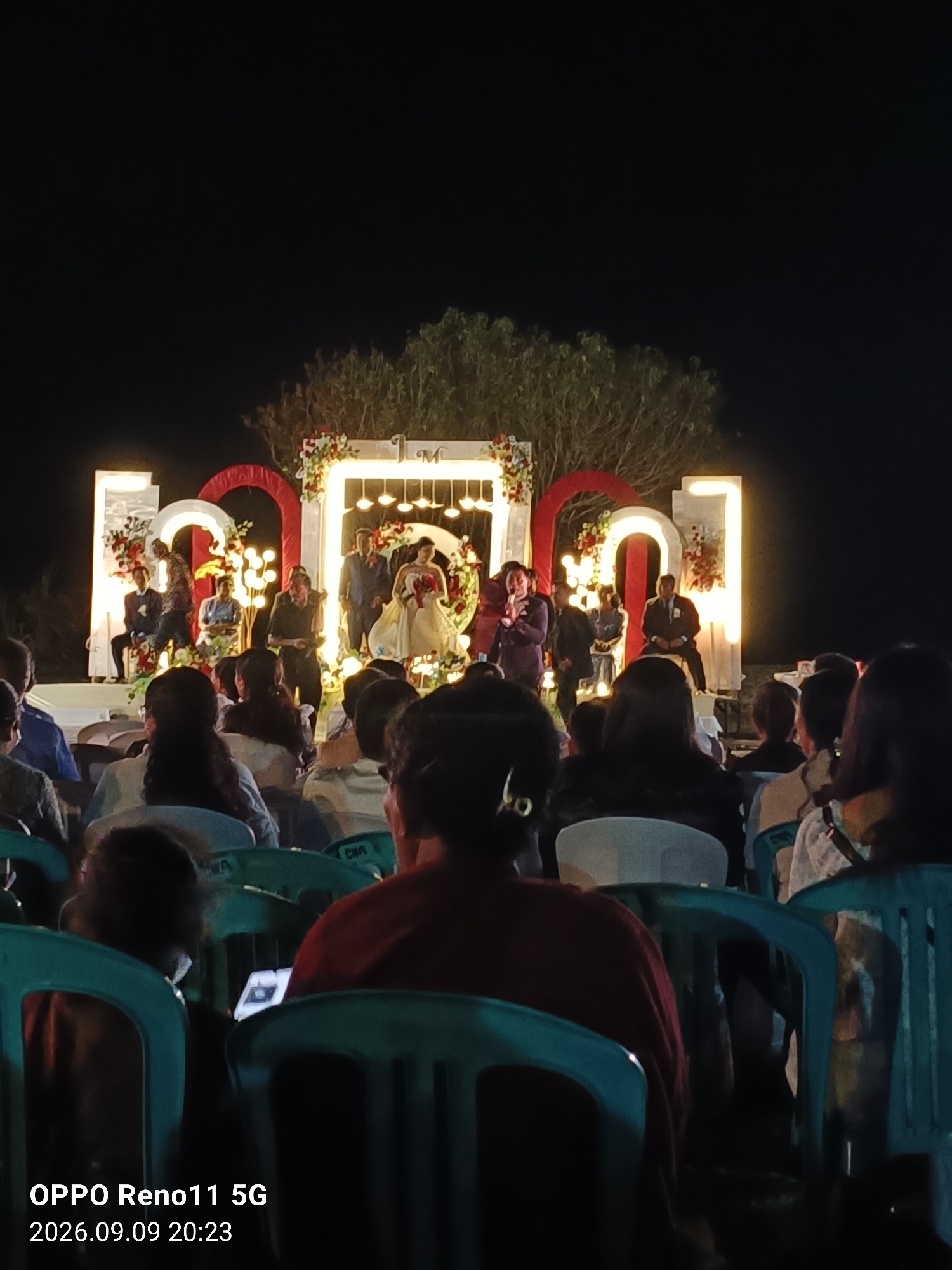 An outdoor wedding reception is taking place at night in Otan, Indonesia. A wedding couple is on a stage with a floral backdrop and illuminated letters that spell out "M + J". The guests are seated in rows of chairs in front of the stage. The lighting is dim, with spotlights illuminating the stage and string lights decorating the backdrop. The overall atmosphere appears festive and intimate.