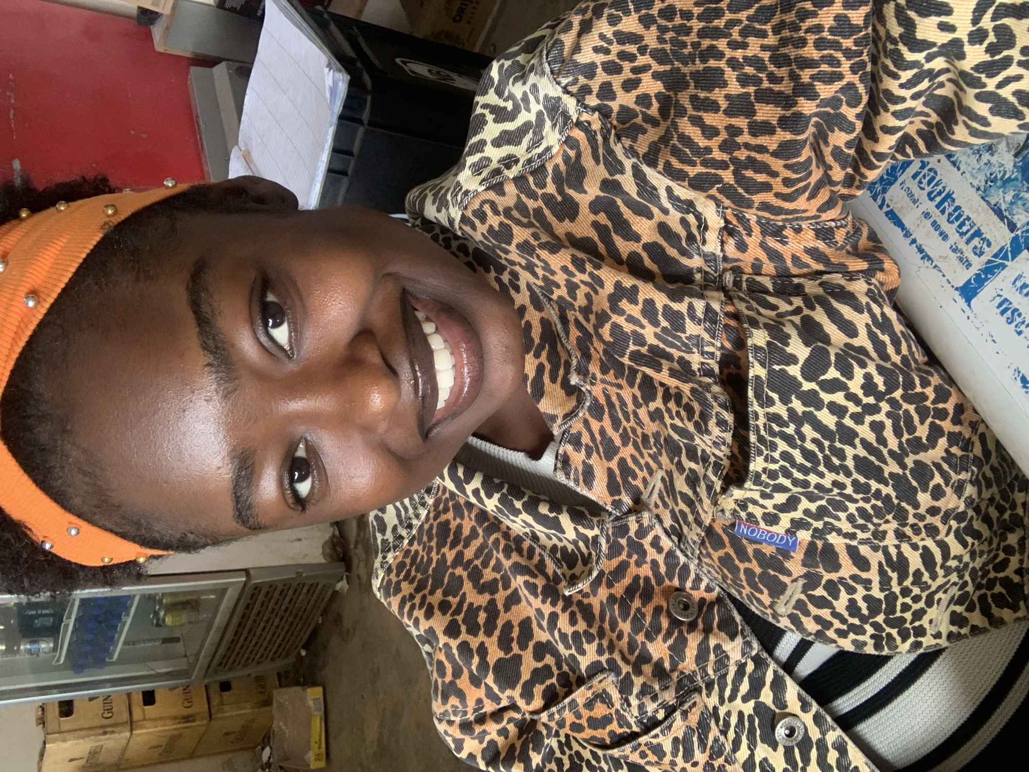 The image shows a close-up of a young Black woman in what appears to be a retail or storage environment in Jos, Nigeria. She is wearing a leopard-print jacket over a striped shirt and an orange headband adorned with silver beads. Her skin is dark and radiant, and she has a bright, wide smile showing her teeth. Her eyes are dark brown and her eyebrows are well-defined. The background suggests a commercial setting; to the left, there are stacked boxes, some labeled "GUINNESS," and a refrigerator filled with drinks. To the right, a black container holds a stack of papers, and a red wall is visible. The lighting is natural, likely from an overhead source, casting soft shadows.