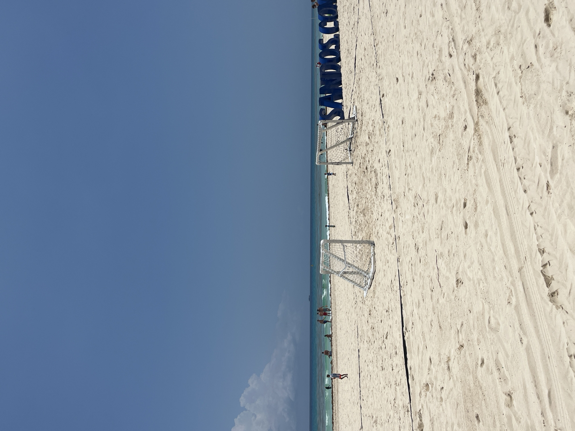 The image is of a beach in Playa del Carmen, Mexico. It shows a bright blue sky, a sandy beach, and the turquoise ocean. There are two small soccer goals set up on the beach. Some people are in the water and walking along the shore. In the background, there is a sign with blue letters that spell out "SANDOS.COM". The sand is light-colored and shows various footprints. There is evidence of vehicles passing along the beach.