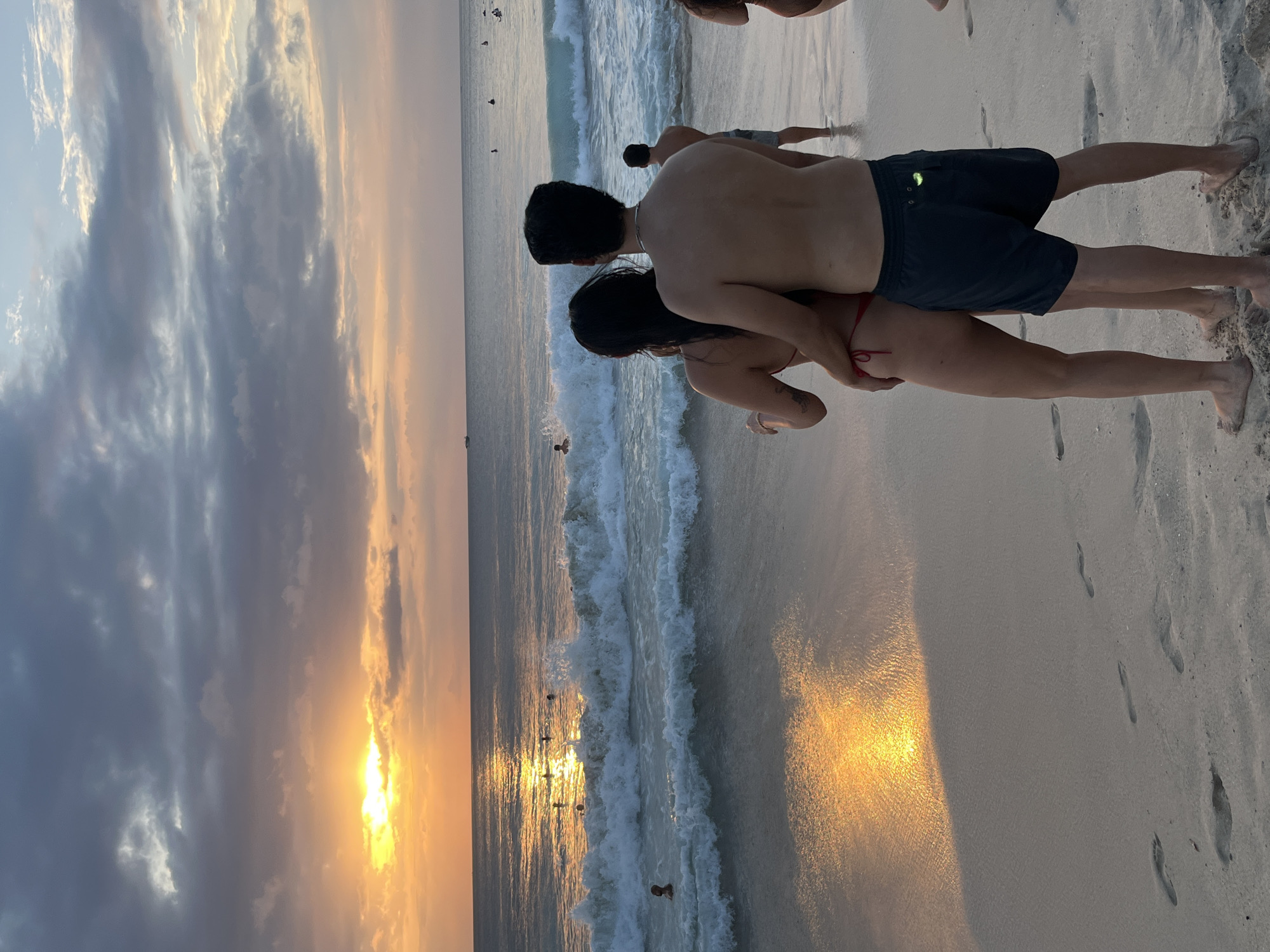 A couple stands on a sandy beach in Kuta Selatan, Indonesia, embracing and facing the ocean at sunset. The sun, partially obscured by clouds, casts a warm glow on the water and the sand, creating reflections. Waves are breaking near the shore, and several other people are visible in the water and on the beach. Footprints are imprinted in the sand in the foreground. The sky is a mix of gray clouds and pastel hues, indicative of the setting sun.