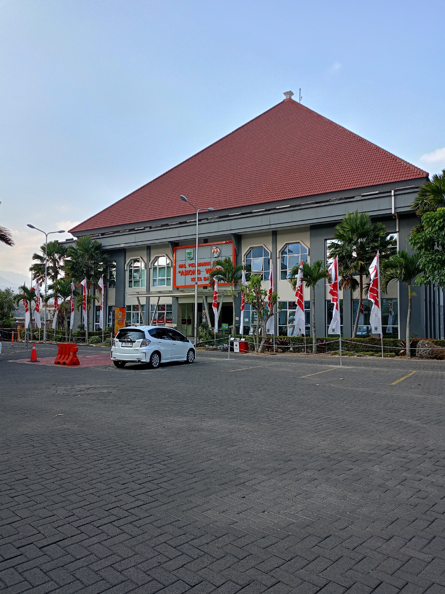 This is a full-shot, eye-level, outdoor photograph taken in the city of Batu, Indonesia, likely during daylight hours under a clear, light blue sky. The main subject is a modern, two-story building with a large, red-tiled, sloped roof. The building's facade is a mix of light gray and dark gray sections, with several large, arched windows featuring tinted glass. An orange banner with text is visible above the entrance.
In front of the building, several Indonesian flags with red and white stripes are planted in the ground, interspersed with palm trees and other greenery. To the left of the flags, there are orange traffic barriers on a paved area. In the foreground, a white hatchback car is parked on a dark gray paved area that appears to be a parking lot. The parking lot is marked with yellow lines. The overall environment suggests a public building or institution.