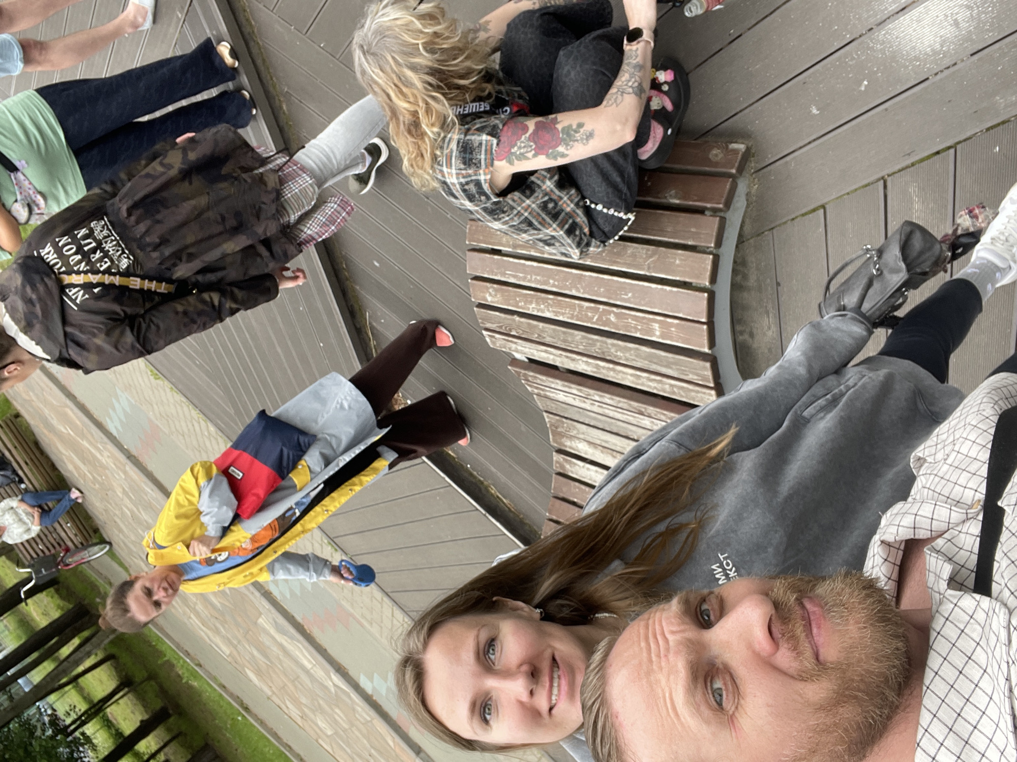 The image shows a group of people in an outdoor setting, possibly a park or a recreational area, as indicated by the wooden decking, benches, and trees visible in the background. The people are in casual attire, and their poses suggest a relaxed and informal gathering. One person is sitting on a bench, wearing a plaid shirt and dark pants, with visible tattoos on their arm. Another person is standing and holding a blue object. Two people are in the foreground, appearing to be taking a selfie. One of them is a woman with long brown hair and a light-colored hoodie, looking at the camera with a smile. The other is a man with a beard and a checkered shirt, also looking towards the camera. There is a colorful jacket with a backpack in the middle ground. A bicycle is partially visible.
The time of day appears to be daytime, with natural lighting. The weather seems mild, and the overall mood is lighthearted and friendly. The setting might be a place for leisure activities.
Visible text includes "NEW YORK LONDON MUNICH BERLIN", "SEWENCHO", and what appears to be Cyrillic characters "МИЛ", on the clothing of some individuals. The location context of Sosnoviy Bor, Russia, suggests a specific geographical area for this scene.