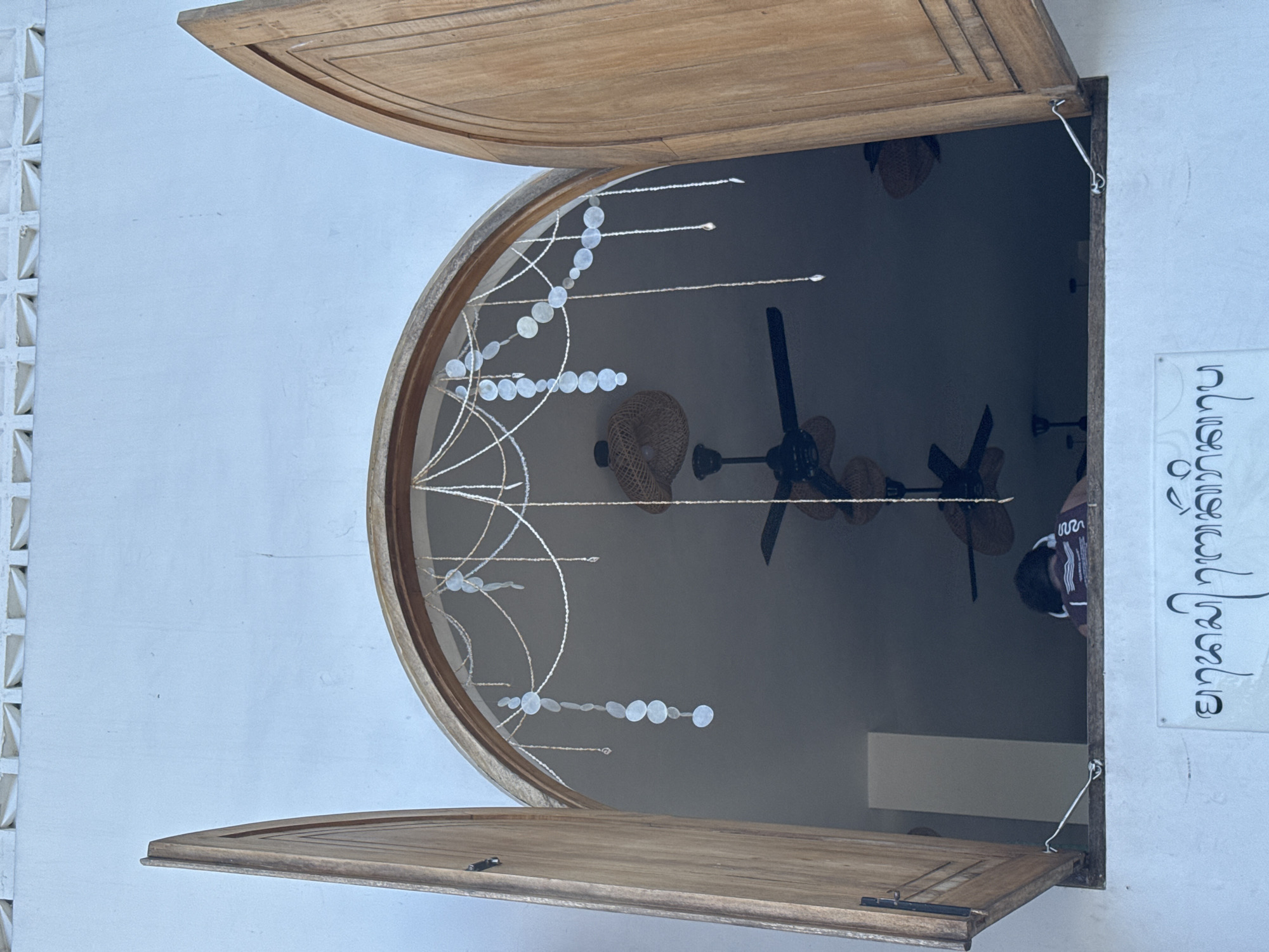 The image shows a window open outward, with a curved top. The window frame and shutters are made of light-colored wood. Inside the window, several ceiling fans are visible in the dark interior. A decorative string of white circles hangs in the window. Outside the window, the wall is white. To the right, a sign with vertically written script is attached to the wall. The scene is located in the city of Kuta Selatan, Indonesia.